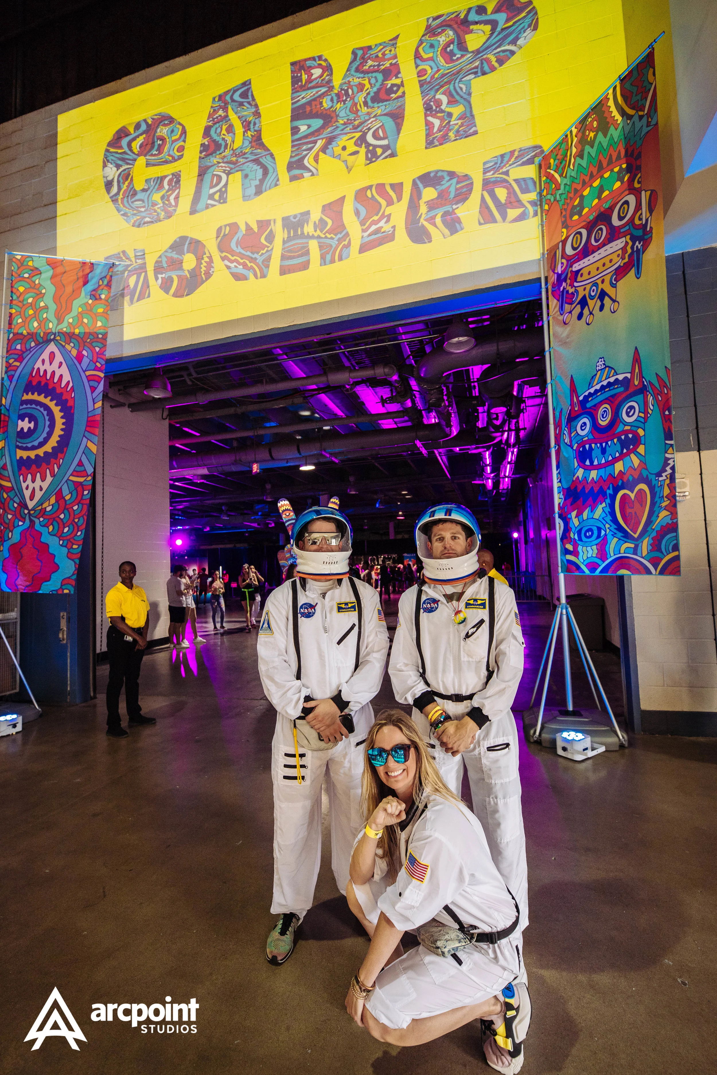Three people dressed as astronauts posing at a vibrant arts or music event with colorful banners and purple lighting.