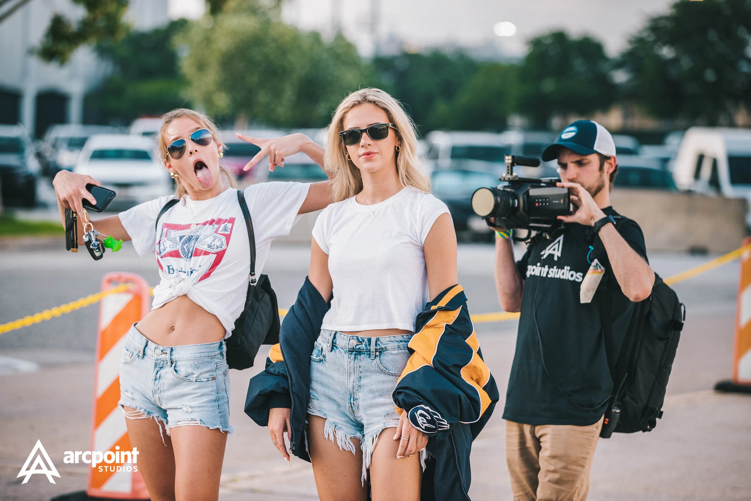 Two young women and a man outdoors near parking lot, one woman sticking out her tongue and making a peace sign, the other standing with arms at her sides, filming crew member with video camera.