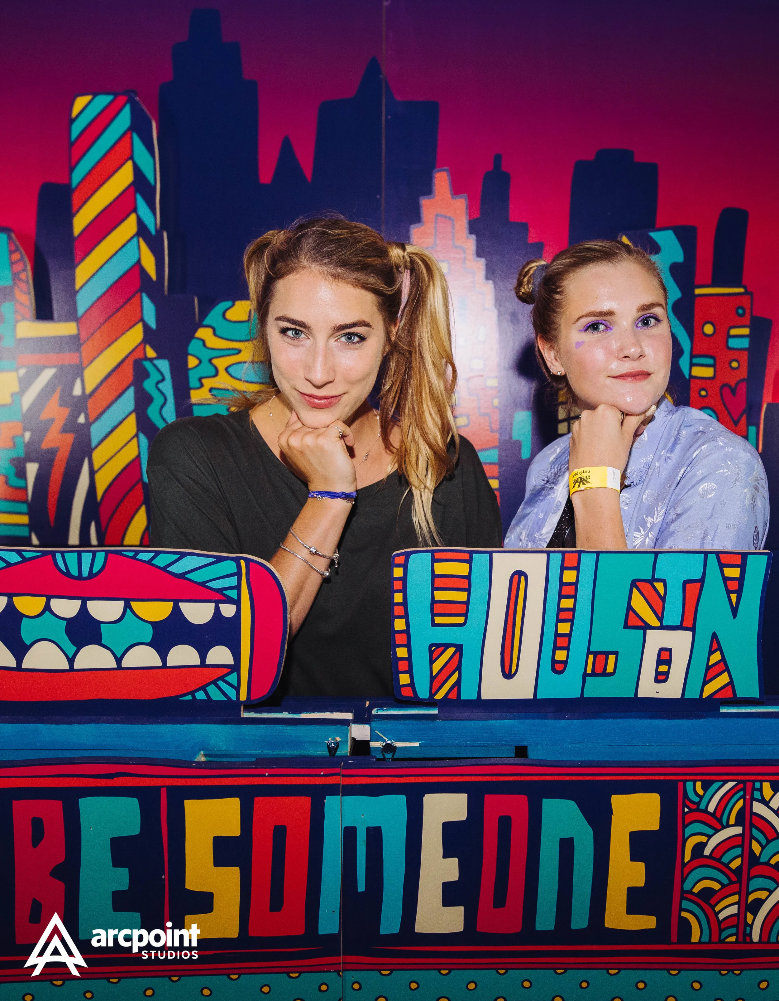 Two women with playful hairstyles and colorful makeup sit behind a vibrant, graffiti-style cityscape backdrop, with signs that read "HOLA!" and "RESUME" in bold, colorful letters.