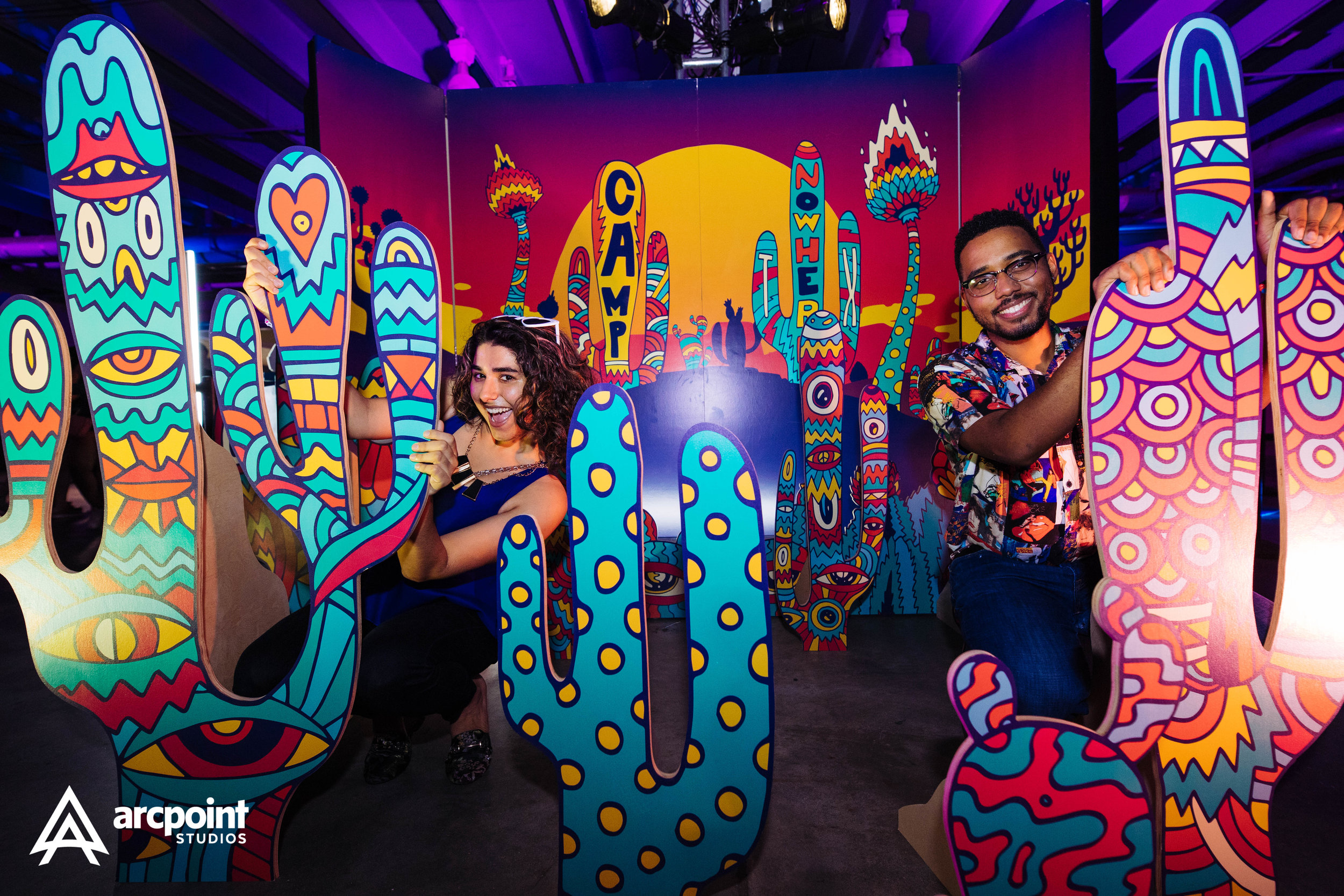 Two smiling people, a woman on the left and a man on the right, play with colorful, cartoonish cactus-shaped cutouts at an art installation with a vibrant desert scene painted backdrop.