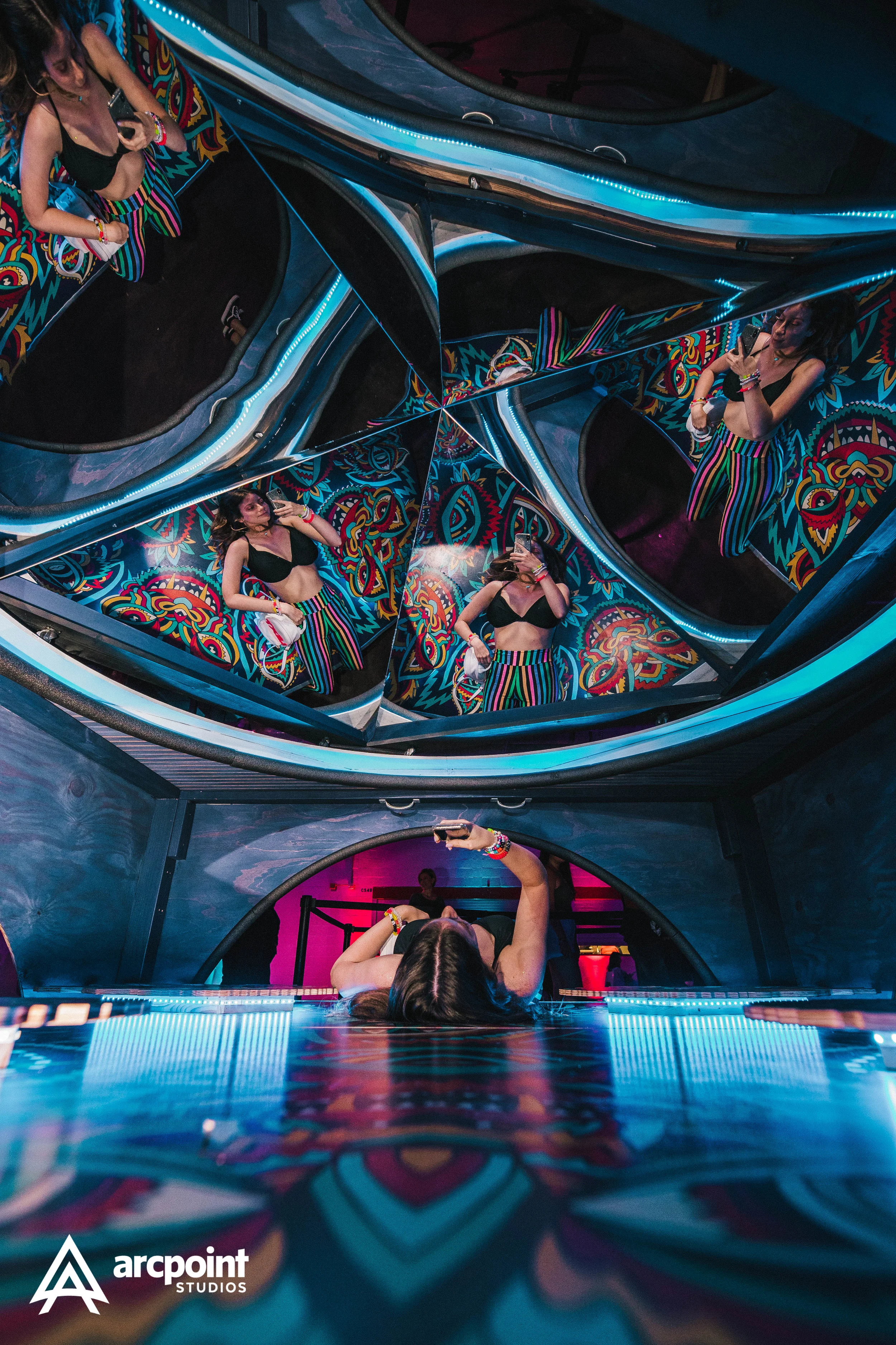 A woman in black crop top and striped pants taking a selfie in a room with kaleidoscopic mirrors and colorful wall art