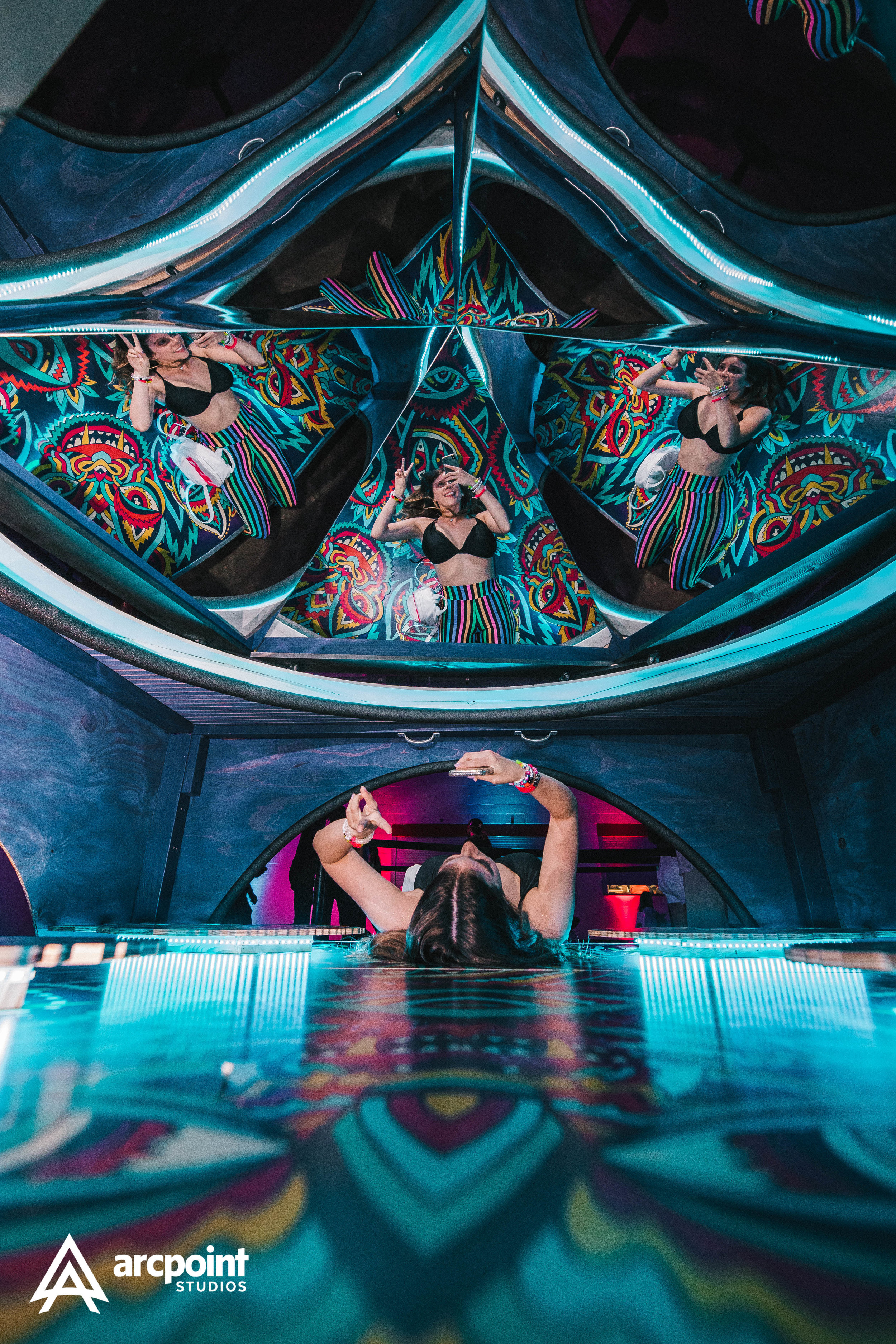 A woman in a black top and striped pants taking a selfie in a multi-level mirror maze with colorful, psychedelic patterned walls under vibrant blue and pink lighting.