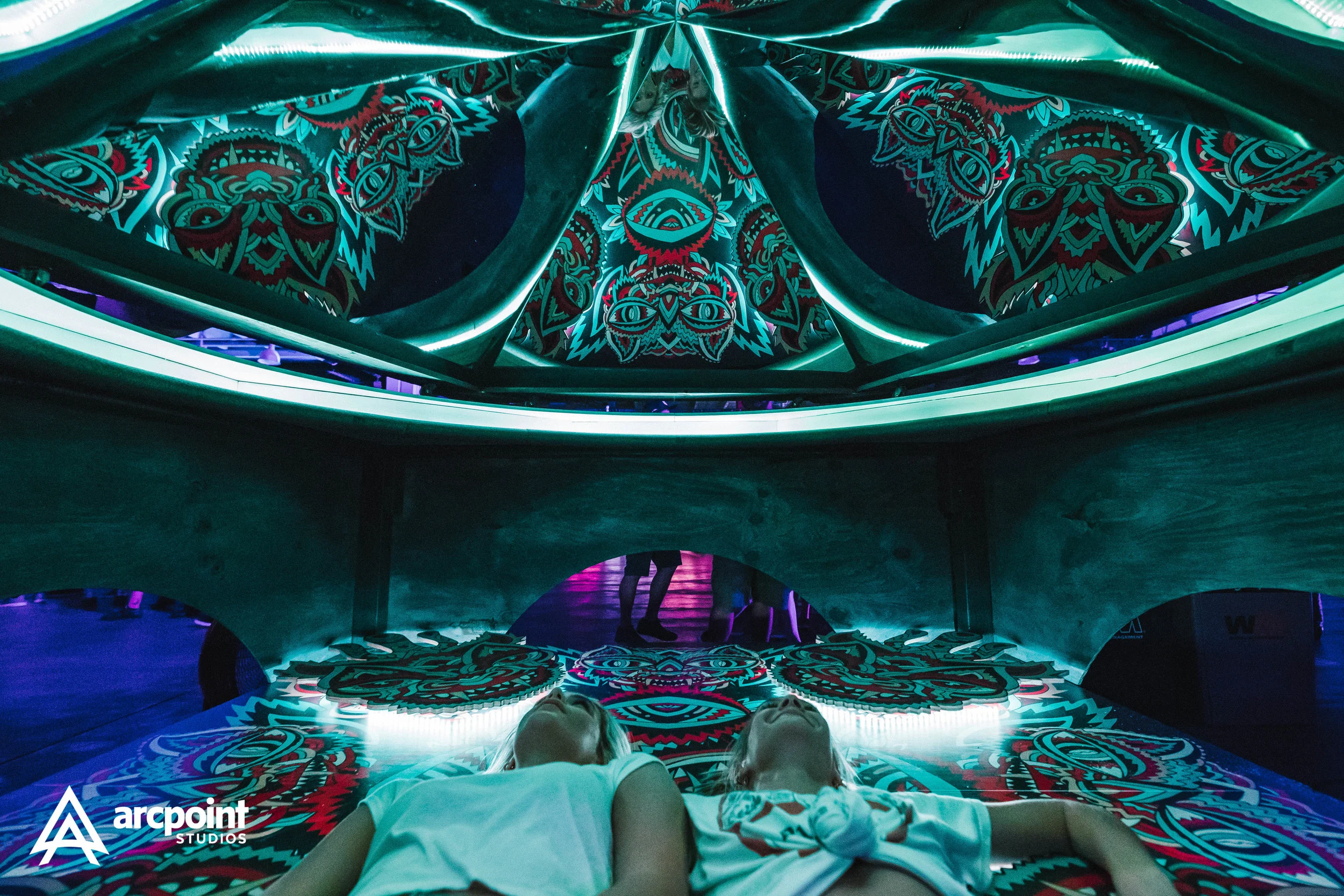 Two children are lying on a psychedelic, brightly colored floor with a mirror ceiling reflecting intricate, symmetrical patterns. The ceiling is illuminated and features a vibrant, tribal-inspired design. The scene appears to be in an art installatio