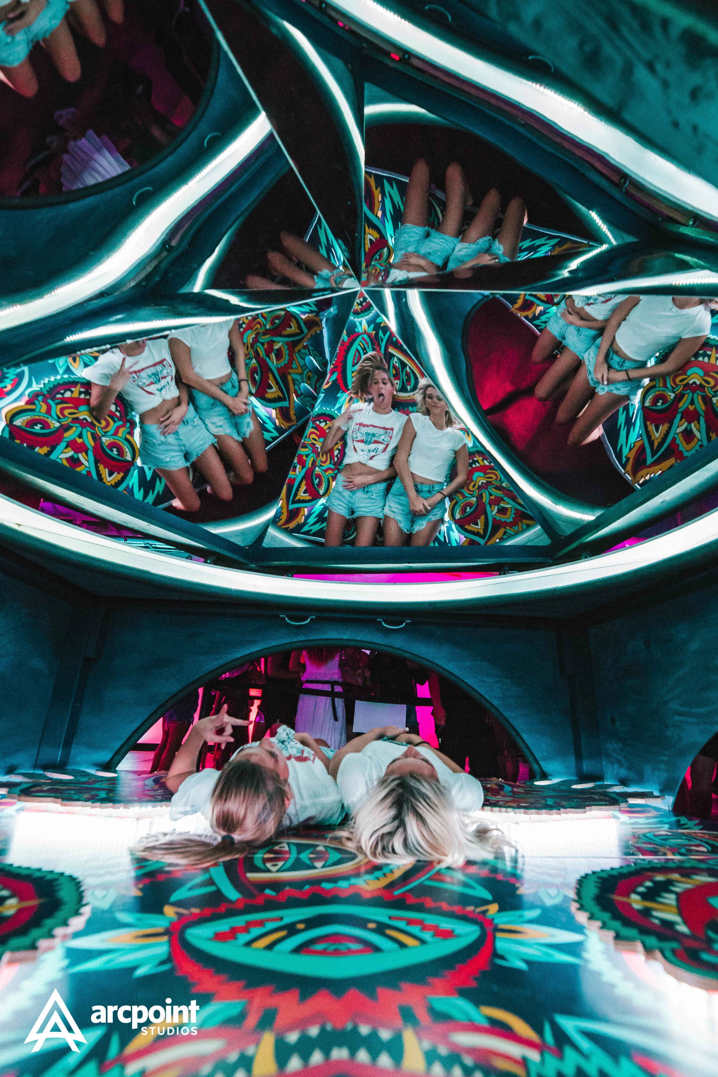People lying in mirrored, kaleidoscope-like chambers with colorful, psychedelic patterns. Several women are lying down, some making peace signs and others lying close together. The environment has vibrant lighting and reflective surfaces.