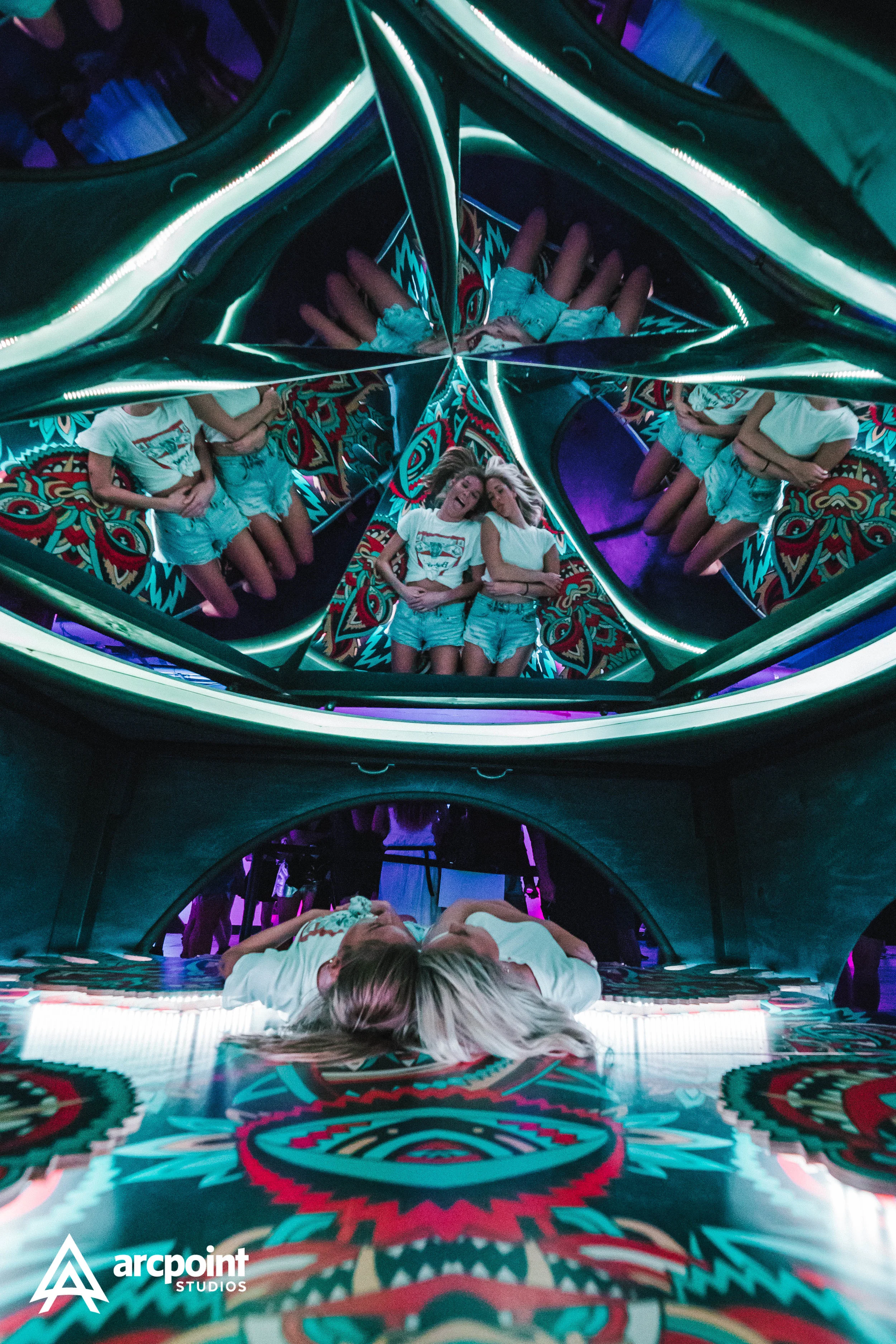 Two girls lying down in a room with colorful patterned walls and mirrors, creating multiple reflections of themselves.