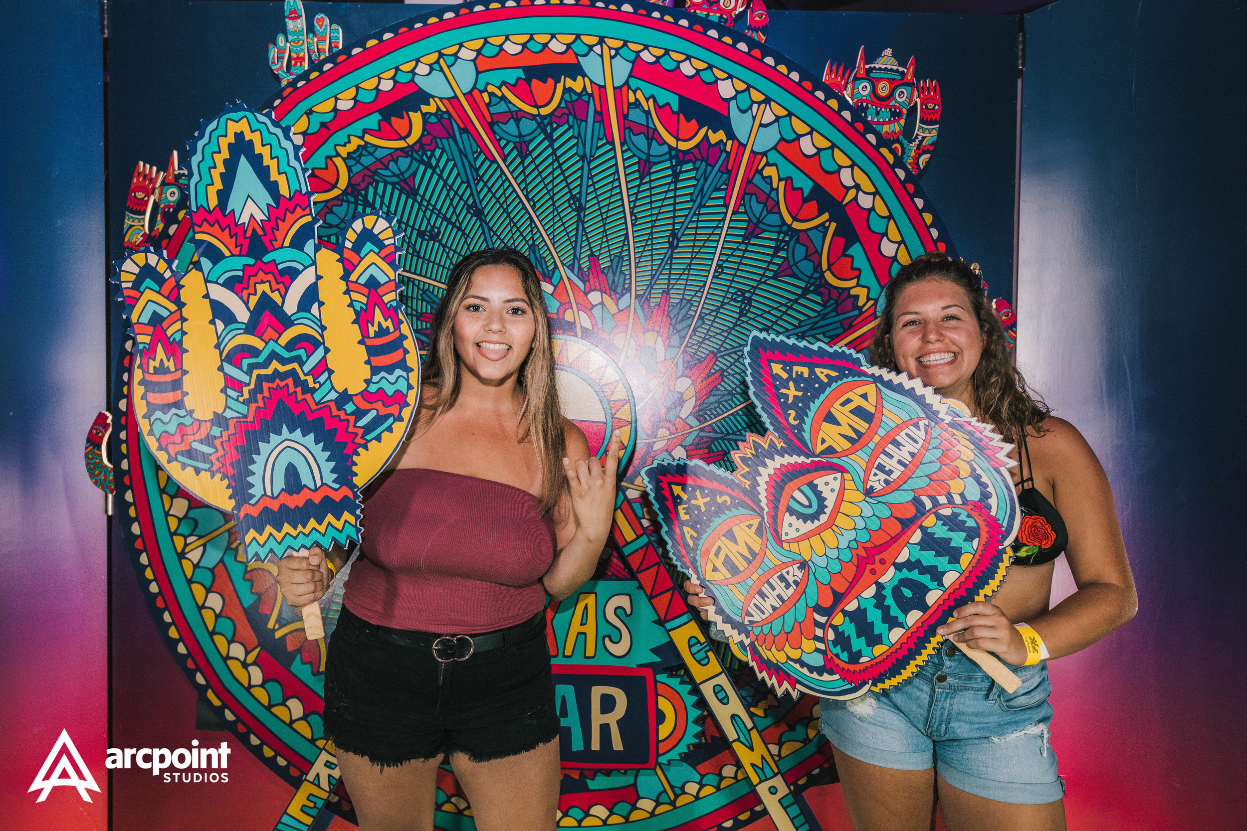 Two young women happily posing with colorful cardboard cutouts in front of a vibrant, intricate background at an event. The woman on the left is wearing a strapless maroon top and black shorts, and the woman on the right is wearing a black top with a