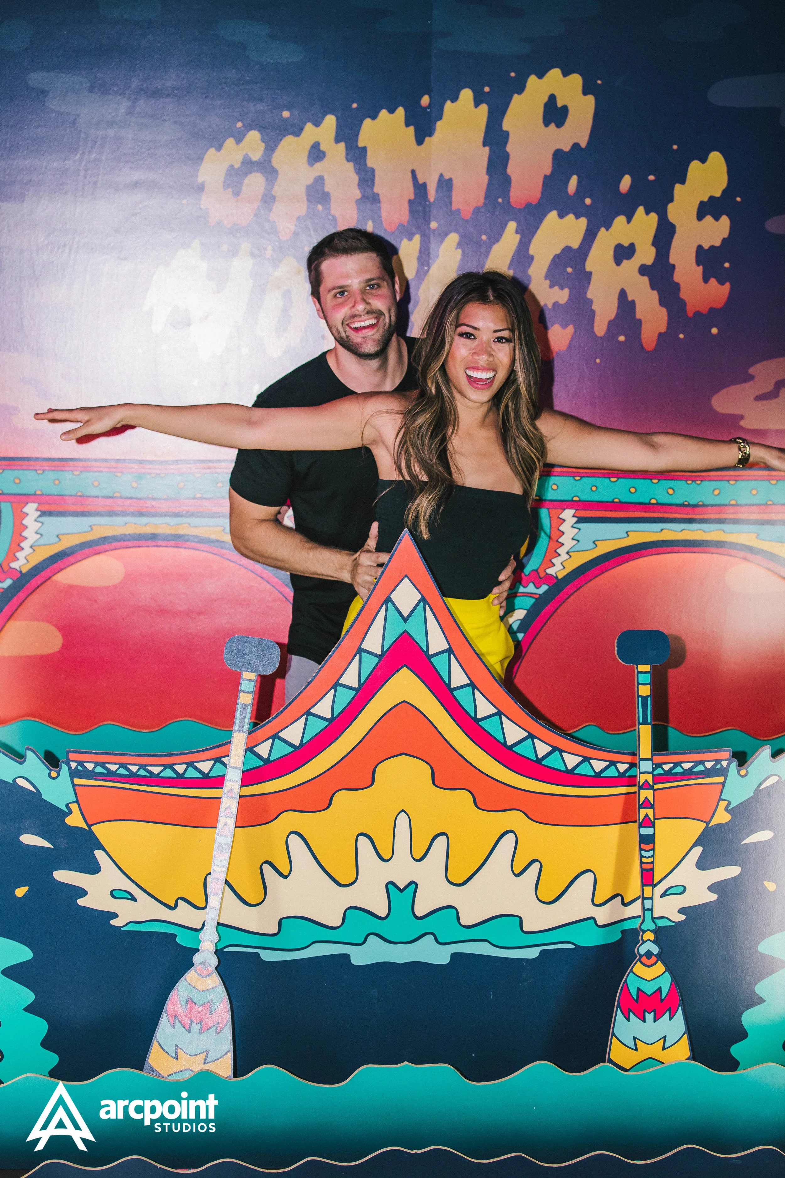 Two people, a man and a woman, smiling and posing with arms extended, behind a colorful cutout of a boat in a fun, vibrant setting at Arcpoint Studios.