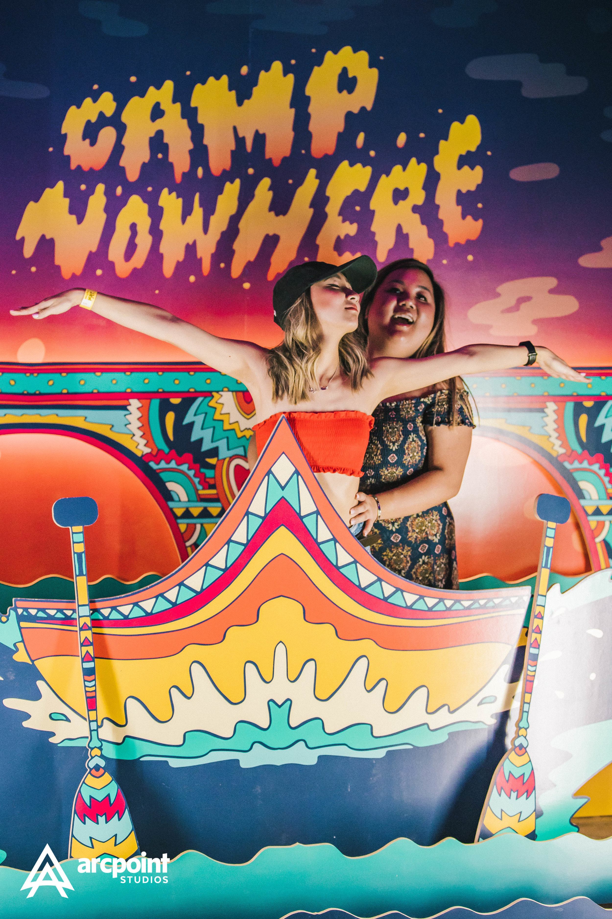 Two women standing in front of a colorful backdrop with the words 'Camp Womance' and a boat illustration. One woman is wearing a black hat and orange top, extending her arms, while the other woman is wearing a floral dress, standing behind her.