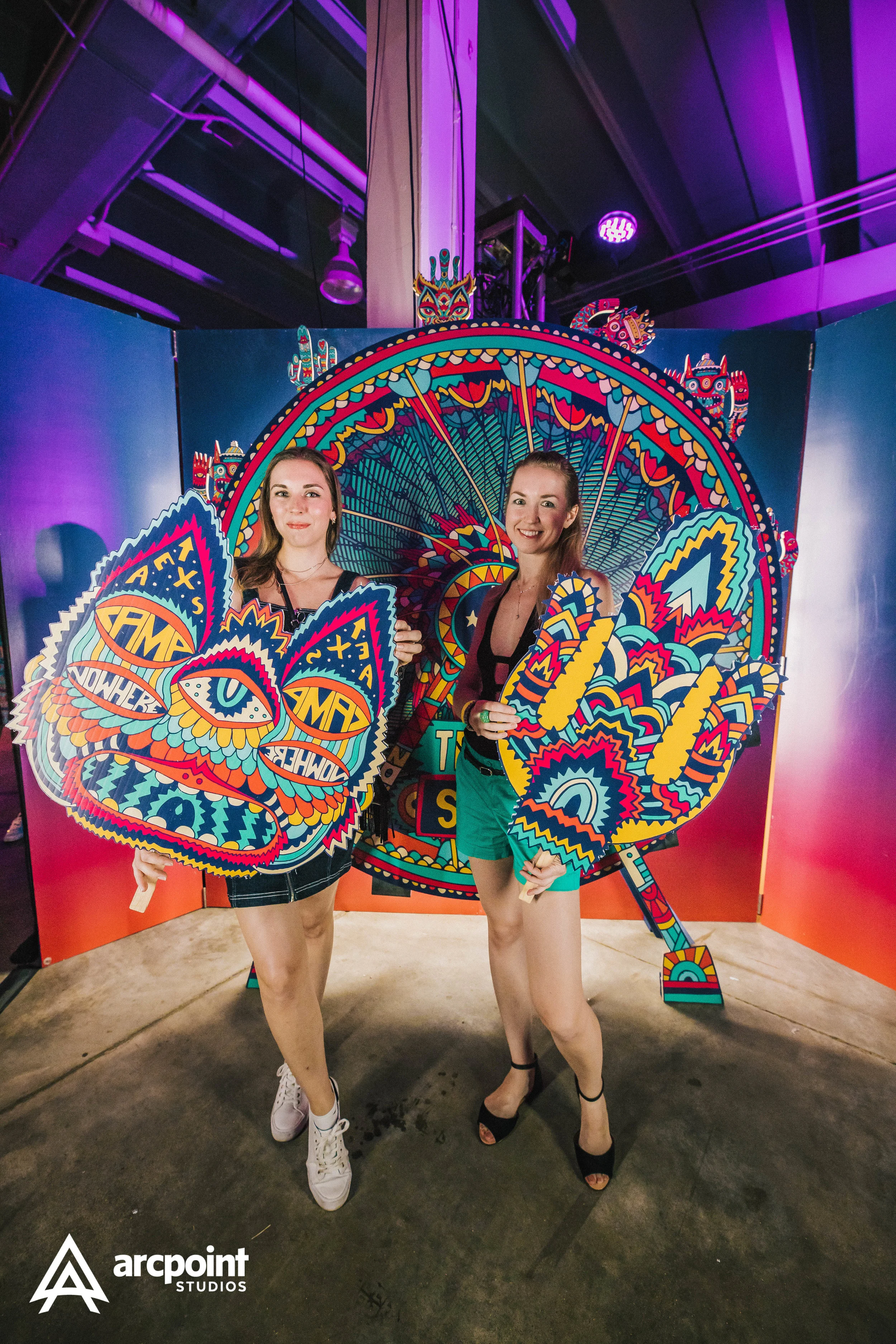 Two women holding colorful carnival masks in front of a vibrant, decorated background with a ferris wheel and geometric patterns, illuminated with purple and pink lighting.