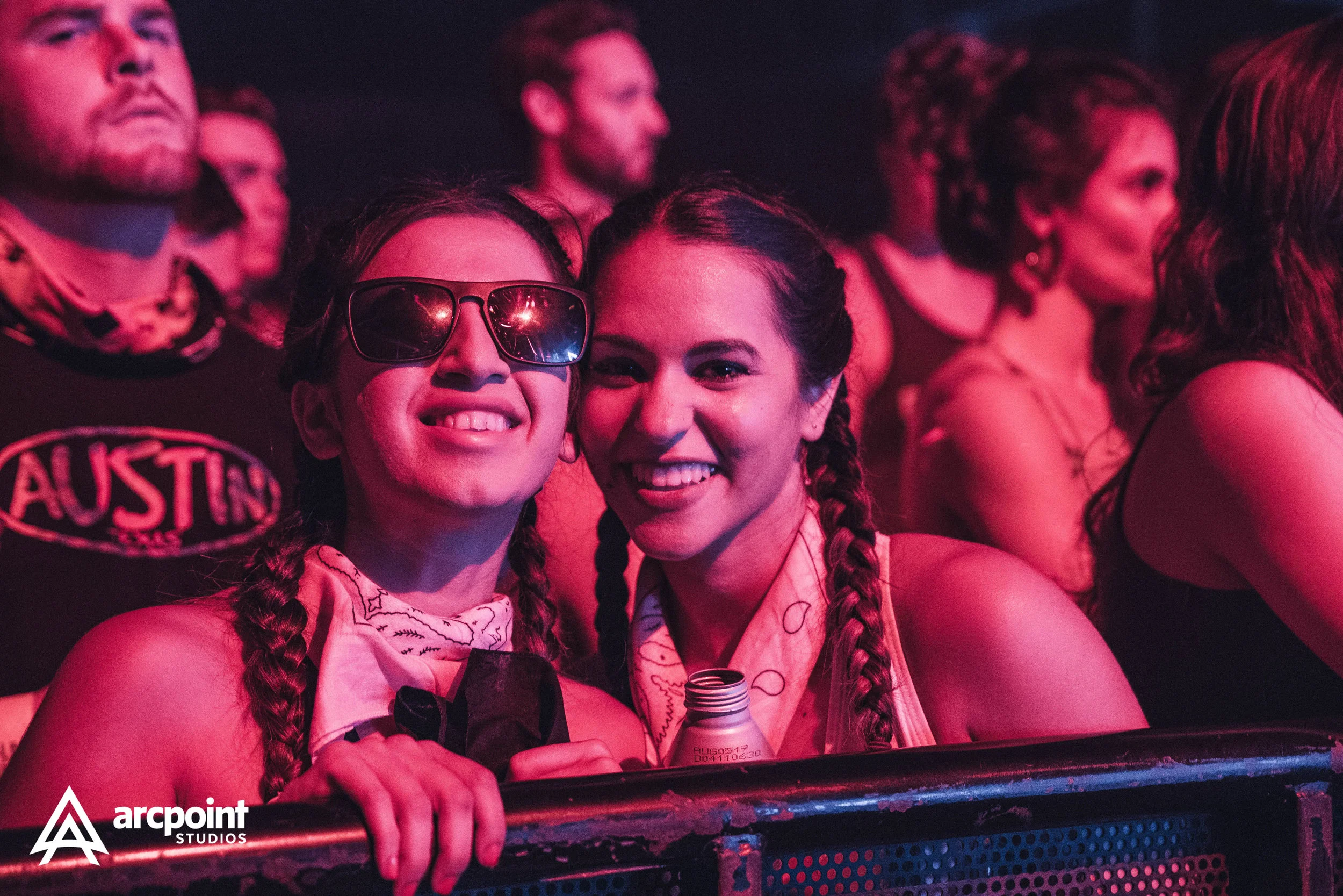 Two young women at a concert, smiling and posing for the camera. One is wearing sunglasses and a bandana, the other has braided hair and is holding a water bottle. There are other people in the background.