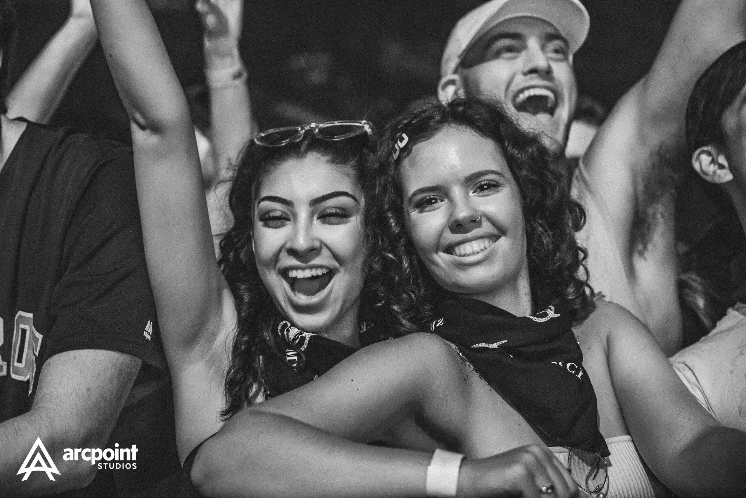 Two young women smiling and enjoying a concert or event, one winking, both wearing bandanas around their necks, with a crowd of people in the background.