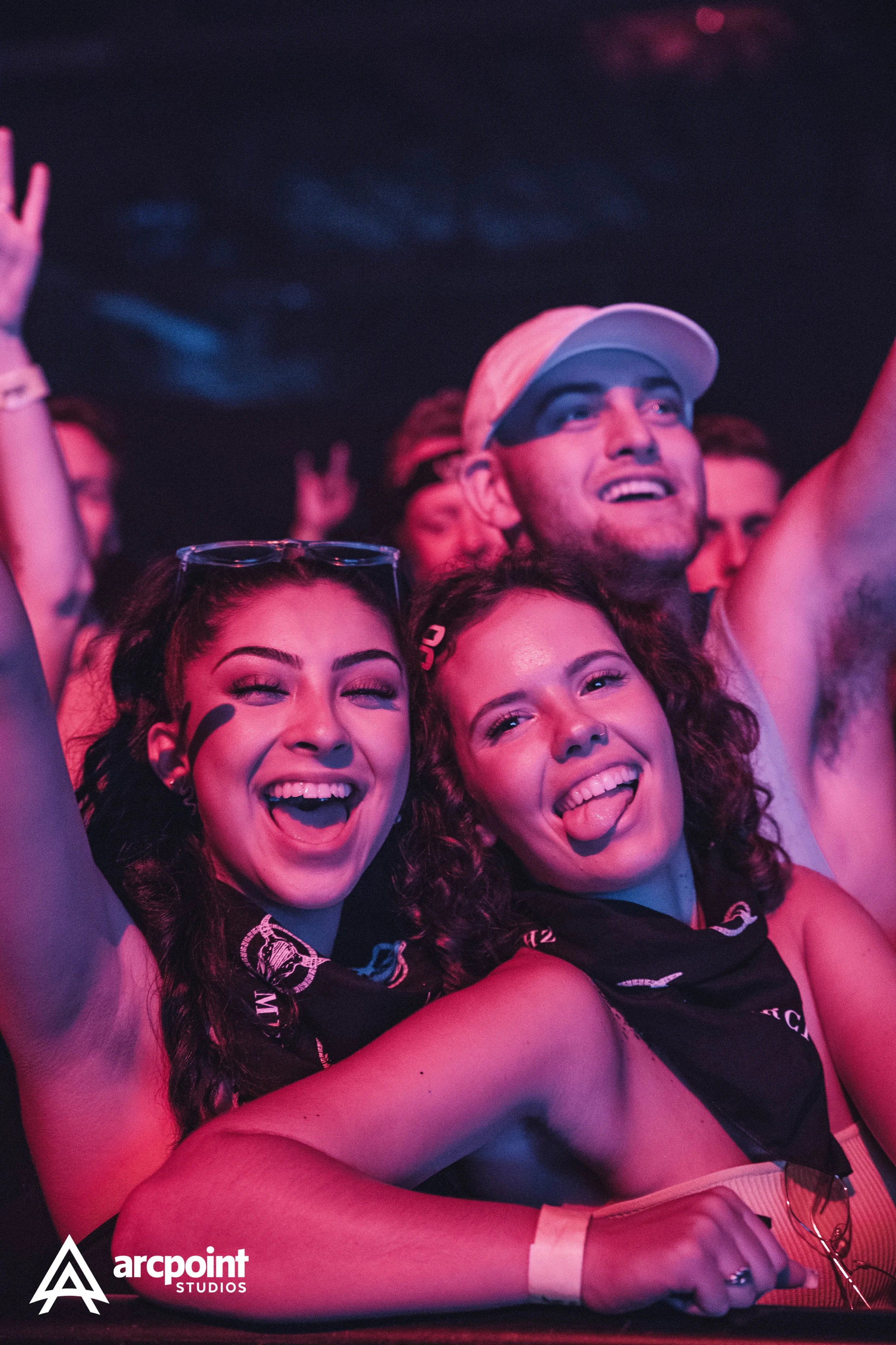 Two women and a man at a concert or festival, smiling and having fun. The women are making excited expressions, with one sticking out her tongue. The man wears a backward cap and a face mask under his eyes.