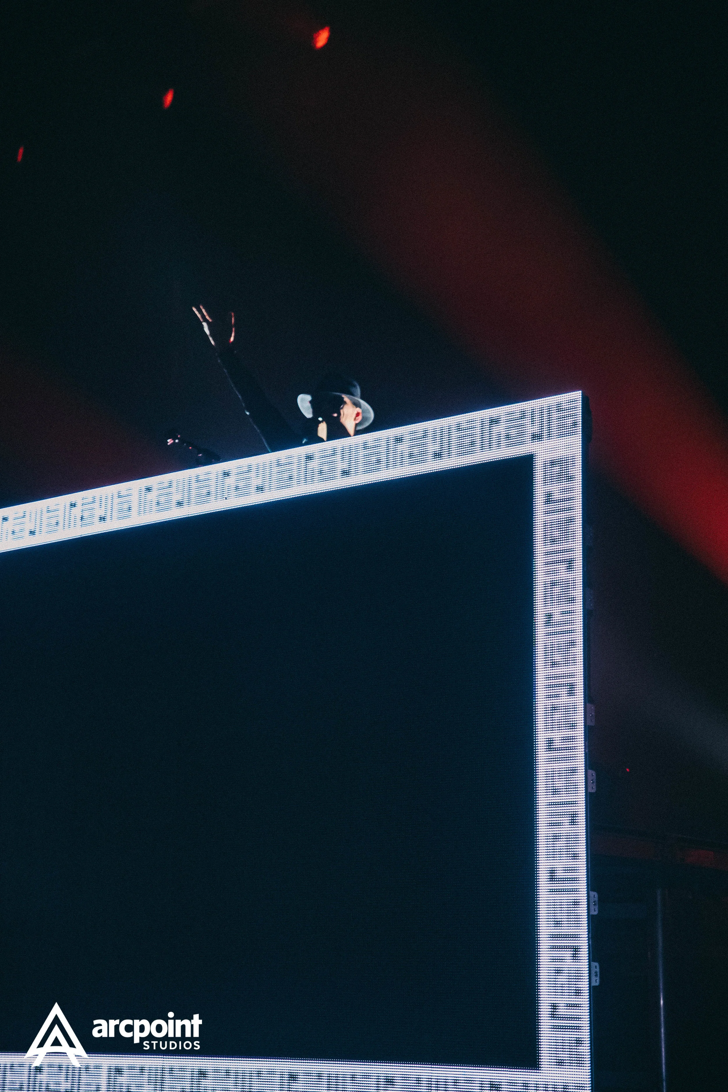 A DJ wearing a black hat and sunglasses is standing behind a large LED screen with the logo 'arcpoint STUDIOS' in the lower left corner, raising one hand in the air during a nighttime event.