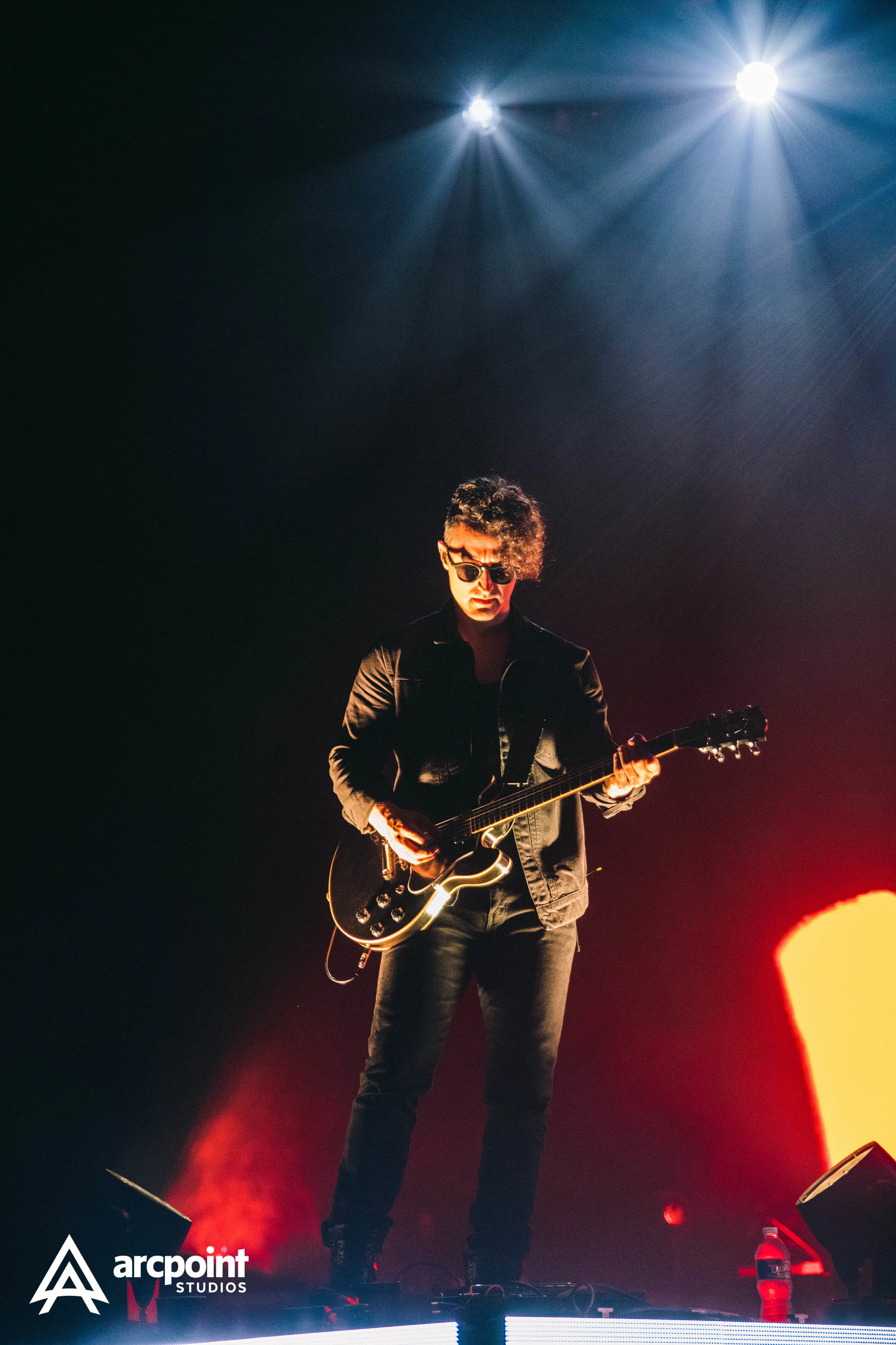 A musician playing an electric guitar on stage, illuminated by bright stage lights, with a dark background and a water bottle on the stage.