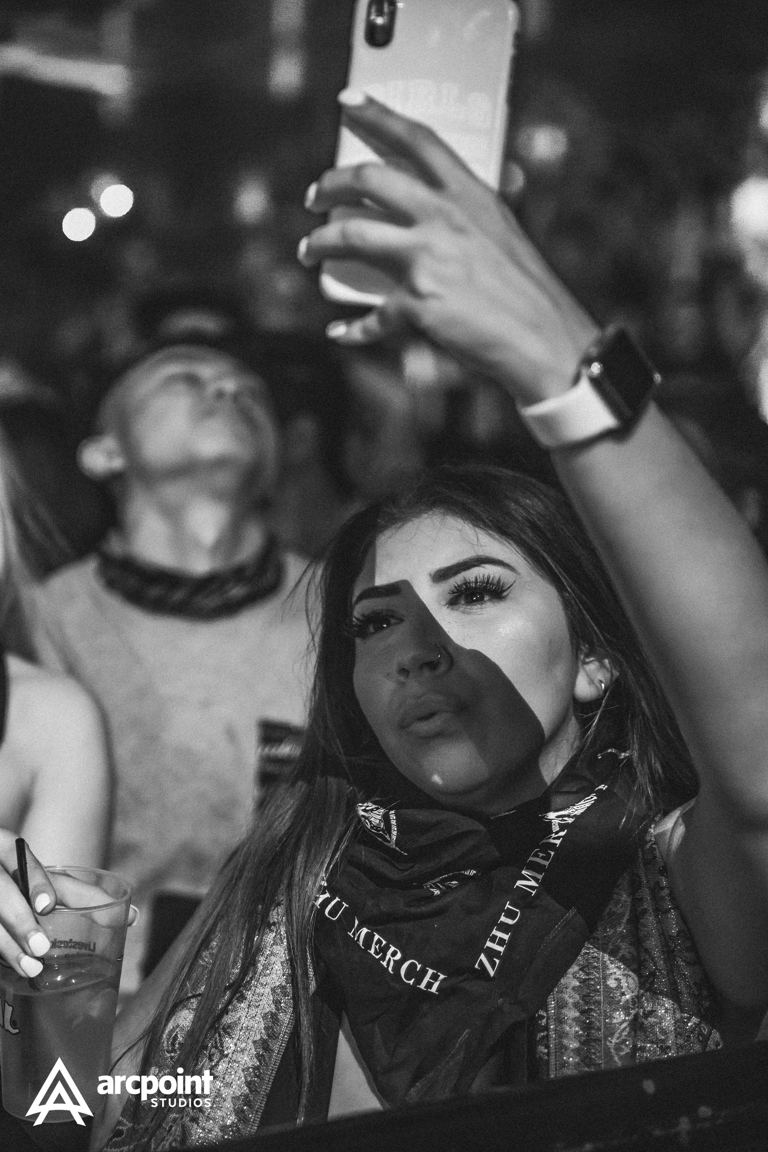 A woman taking a selfie at a crowded event, with a shadow cast across her face, and a man in the background raising his hand.