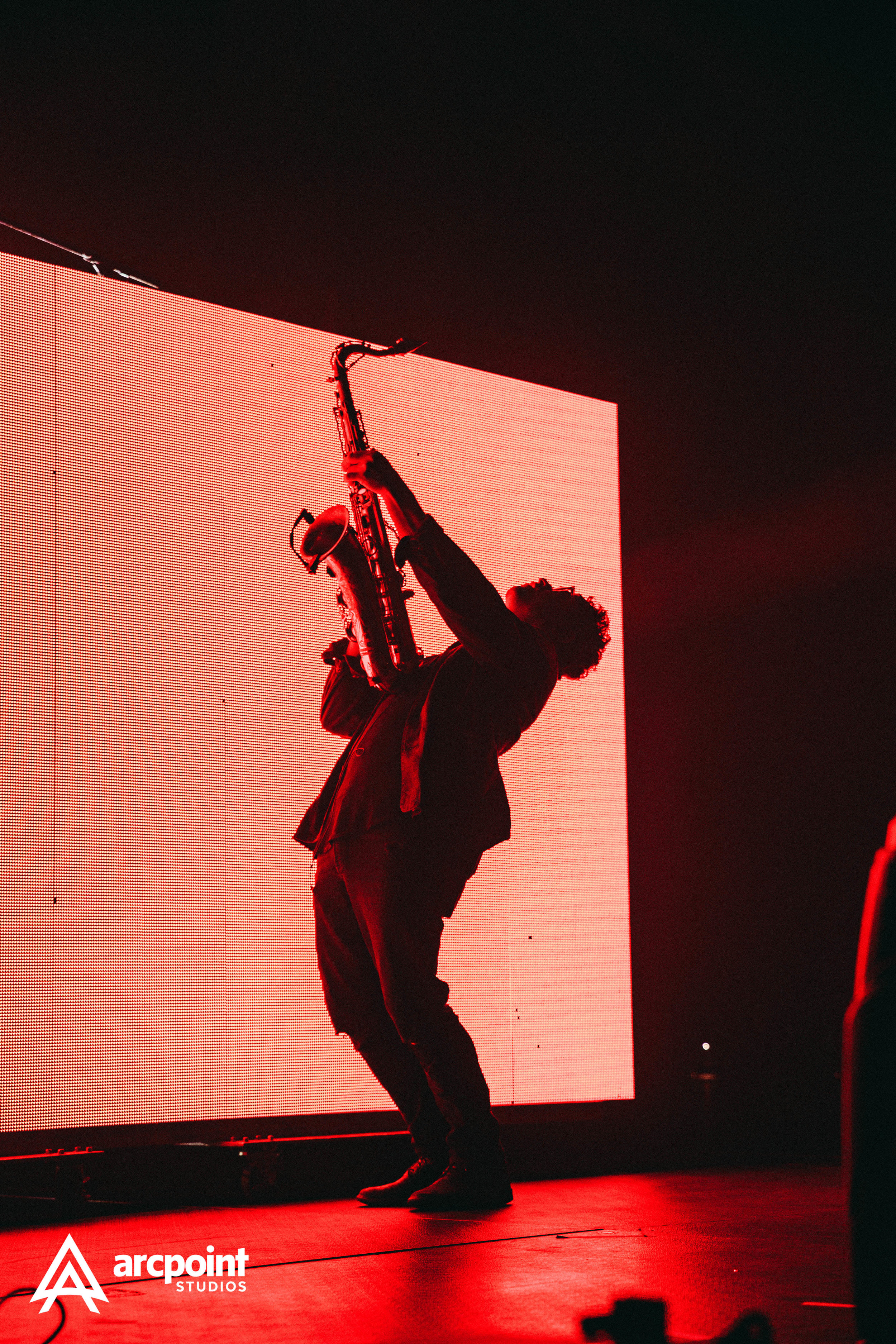 A musician wearing dark clothing and a fedora hat playing a saxophone on stage with a red background