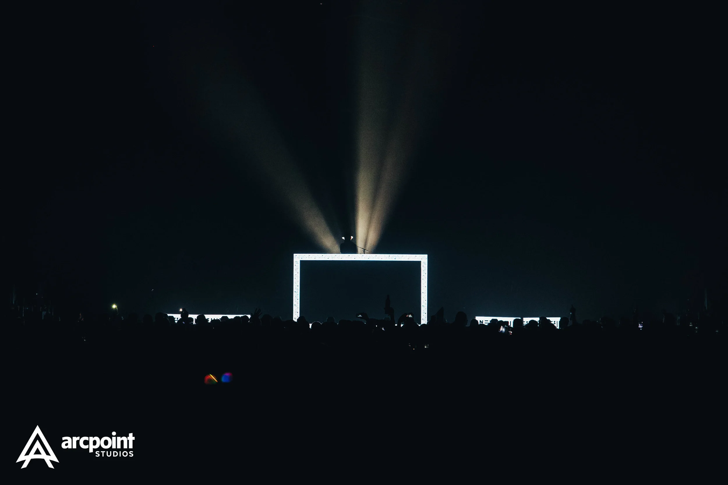 A dark concert scene with a crowd watching a performer on stage, which is illuminated by a rectangular frame with bright lights. The audience is holding up phones, and spotlights are shining down from above.