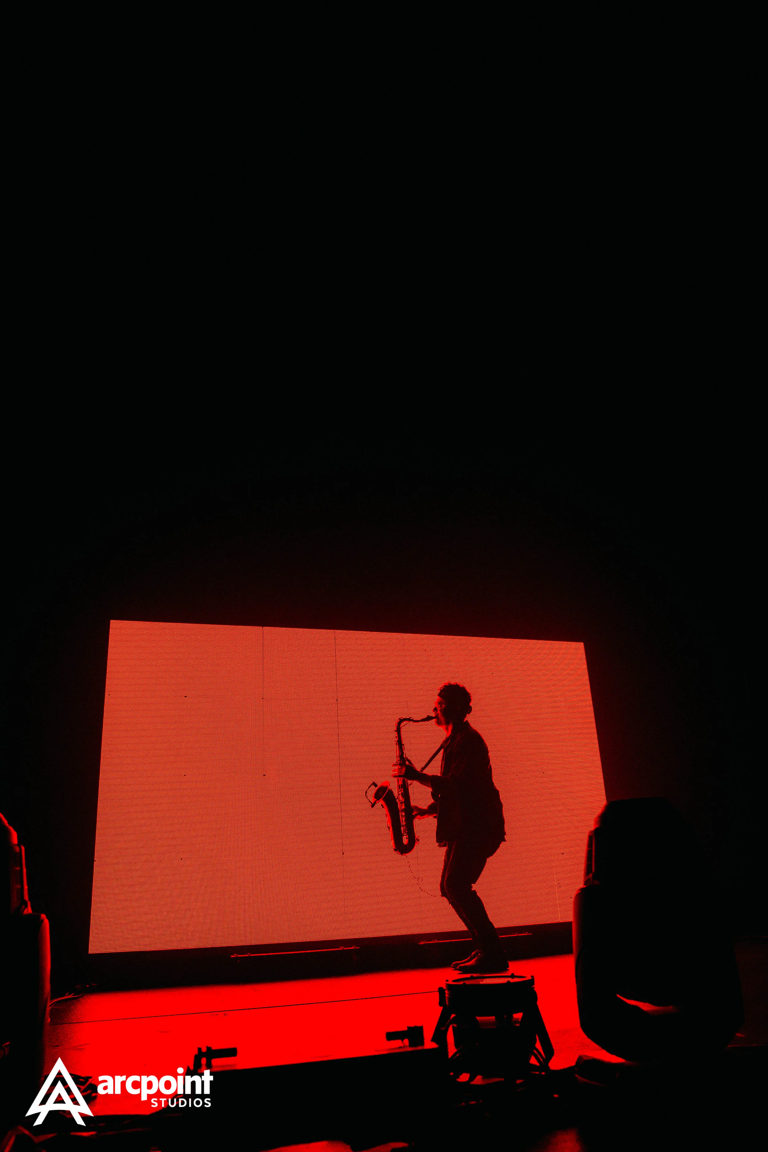 A musician playing saxophone on stage with a red-lit background and LED screen, with the logo 'arcpoint STUDIOS' in the bottom left corner.