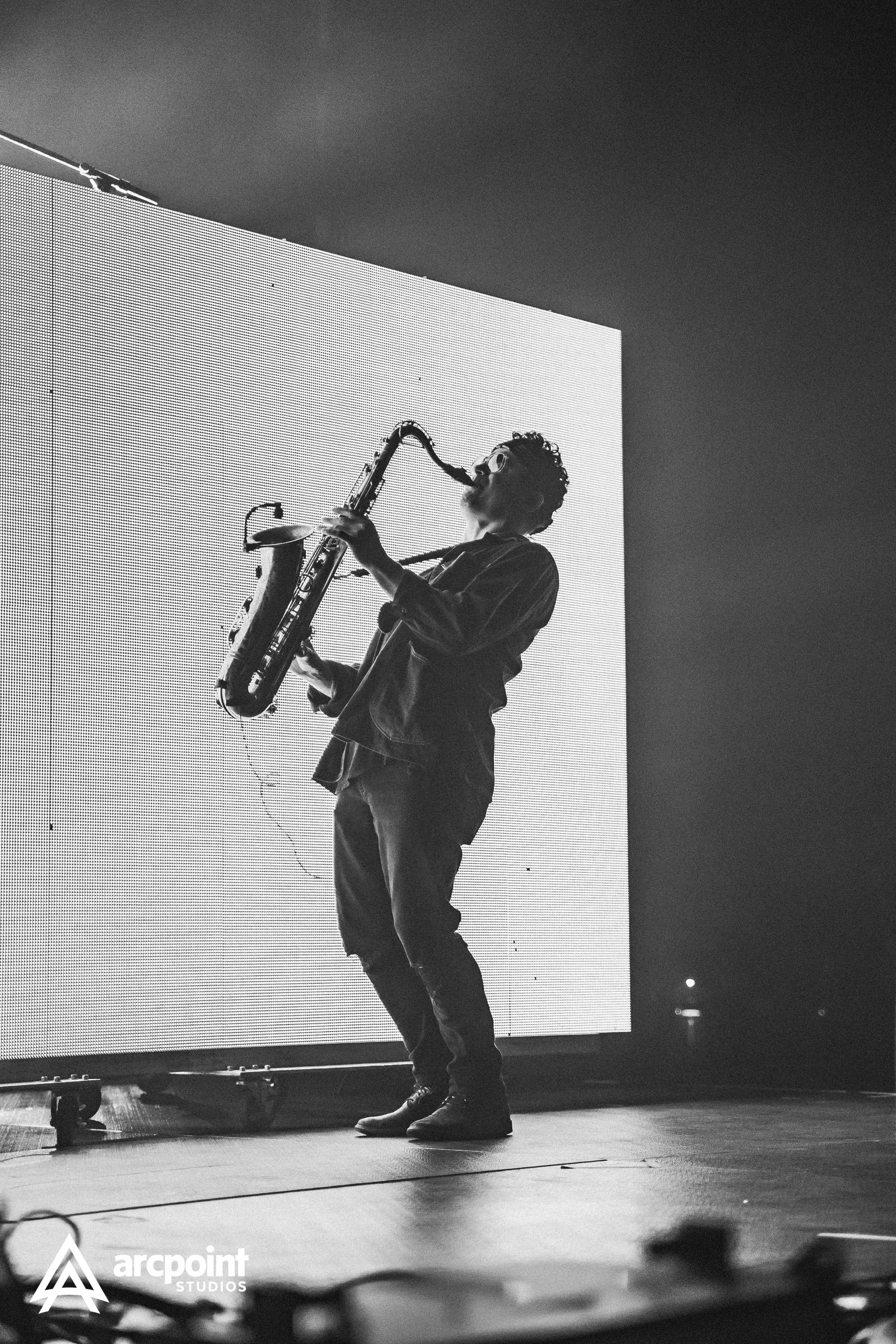 A musician playing a saxophone on stage, with a large LED screen behind him showing his silhouette in black and white.