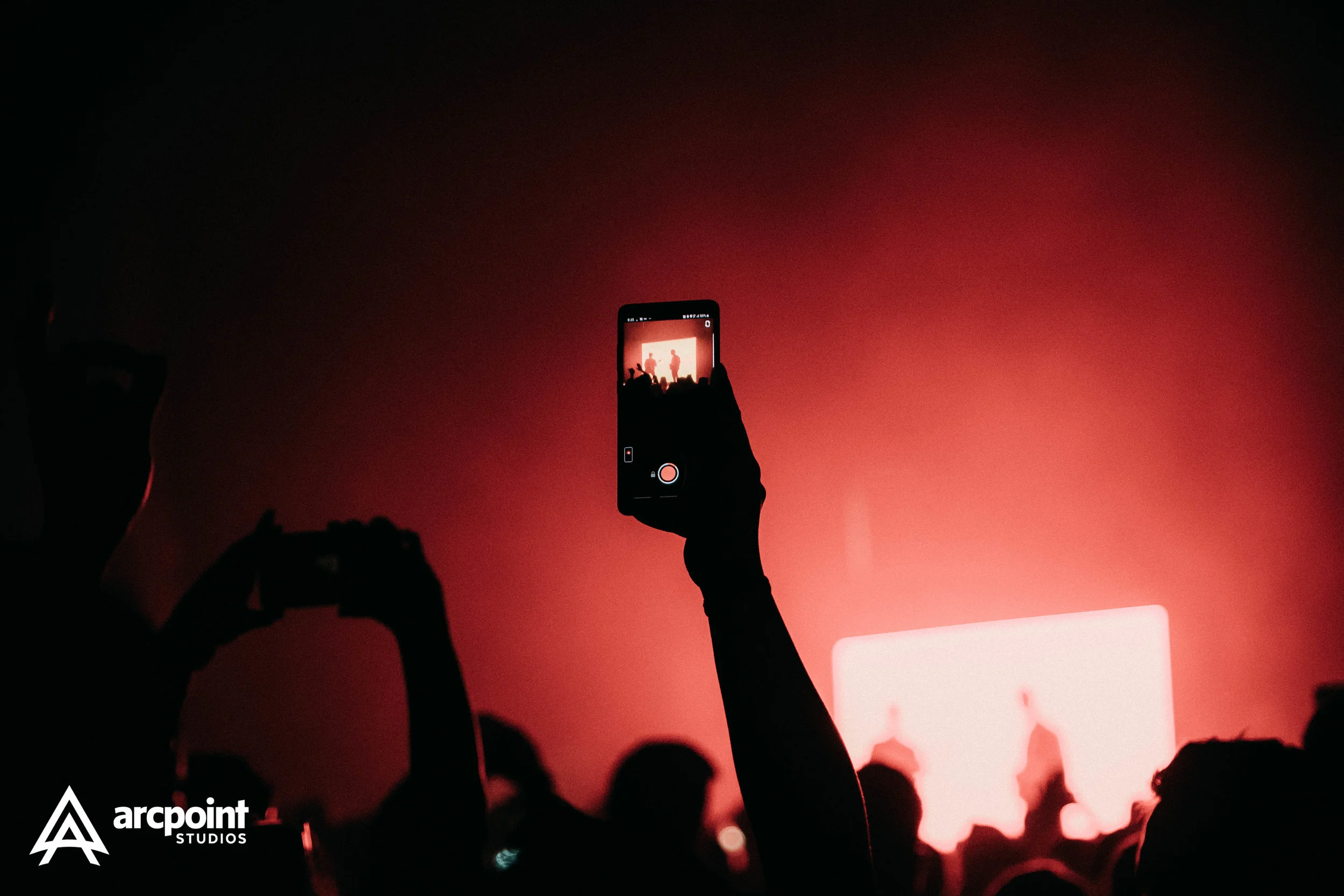 People at a concert or event taking photos of the stage with their smartphones, with red lighting and silhouettes of the crowd.