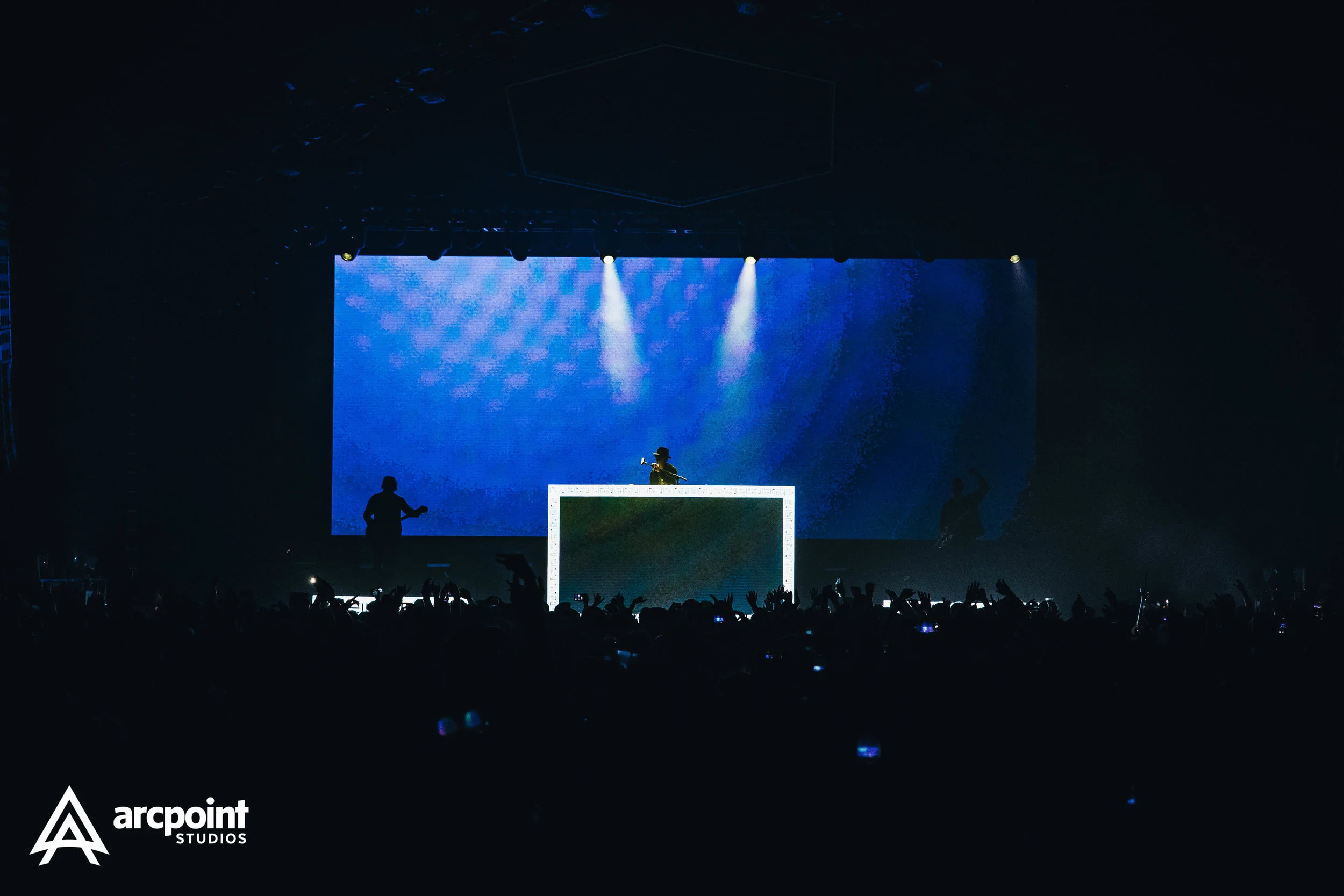 A concert scene with a DJ on stage behind a illuminated DJ booth, silhouetted band members on either side, and a large LED screen in the background displaying blue visuals. The audience is visible in the foreground with raised hands and phones.”}}#en