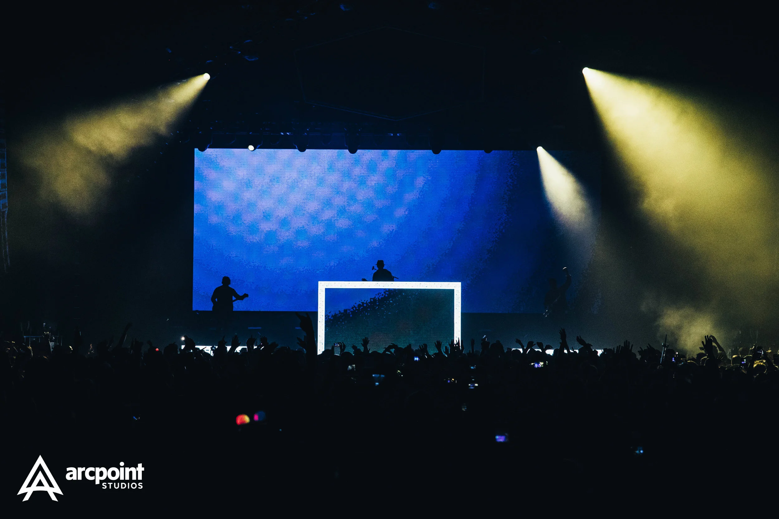 A live concert scene with a DJ on stage, silhouetted against a large LED screen and stage lights, with an audience raising their hands.