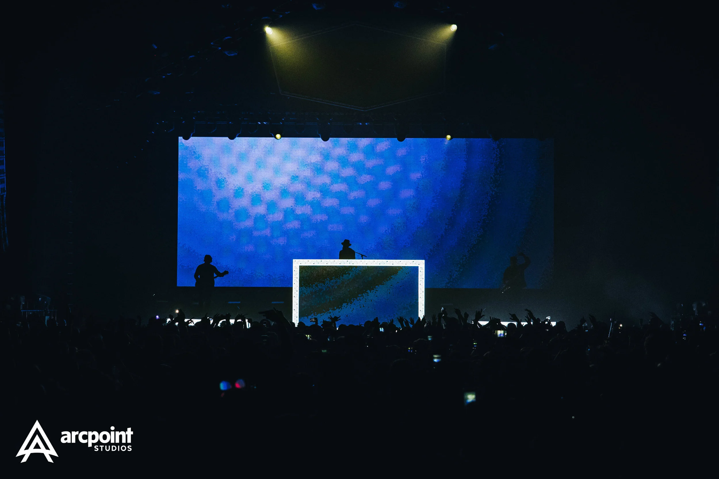 Silhouettes of three performers on stage with a large blue digital backdrop at a concert, with audience hands raised, and 