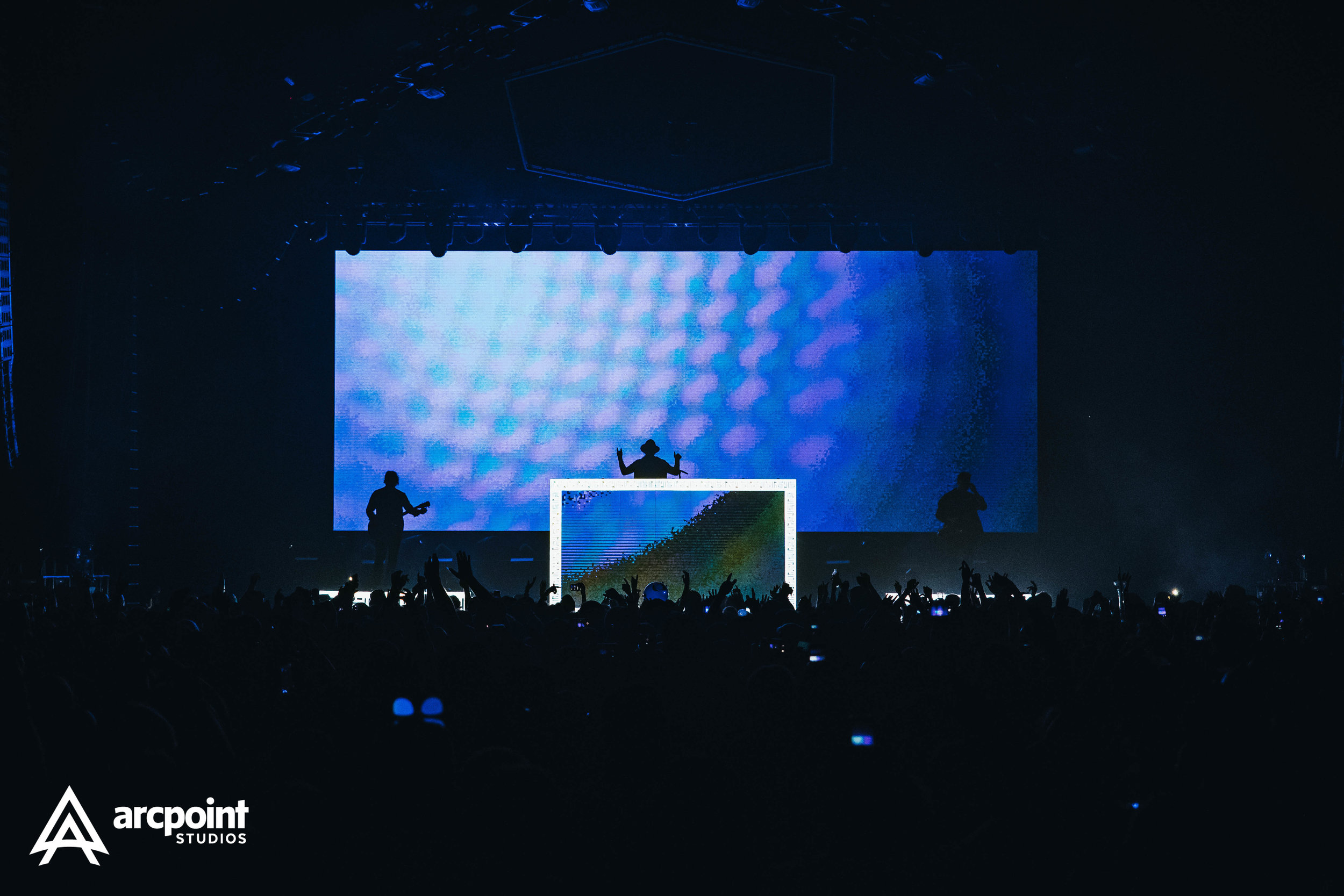 A concert stage with three silhouettes of performers and a large LED screen in the background displaying blue patterns, with an audience in front raising hands and taking photos. The logo of Arcpoint Studios is in the lower left corner.