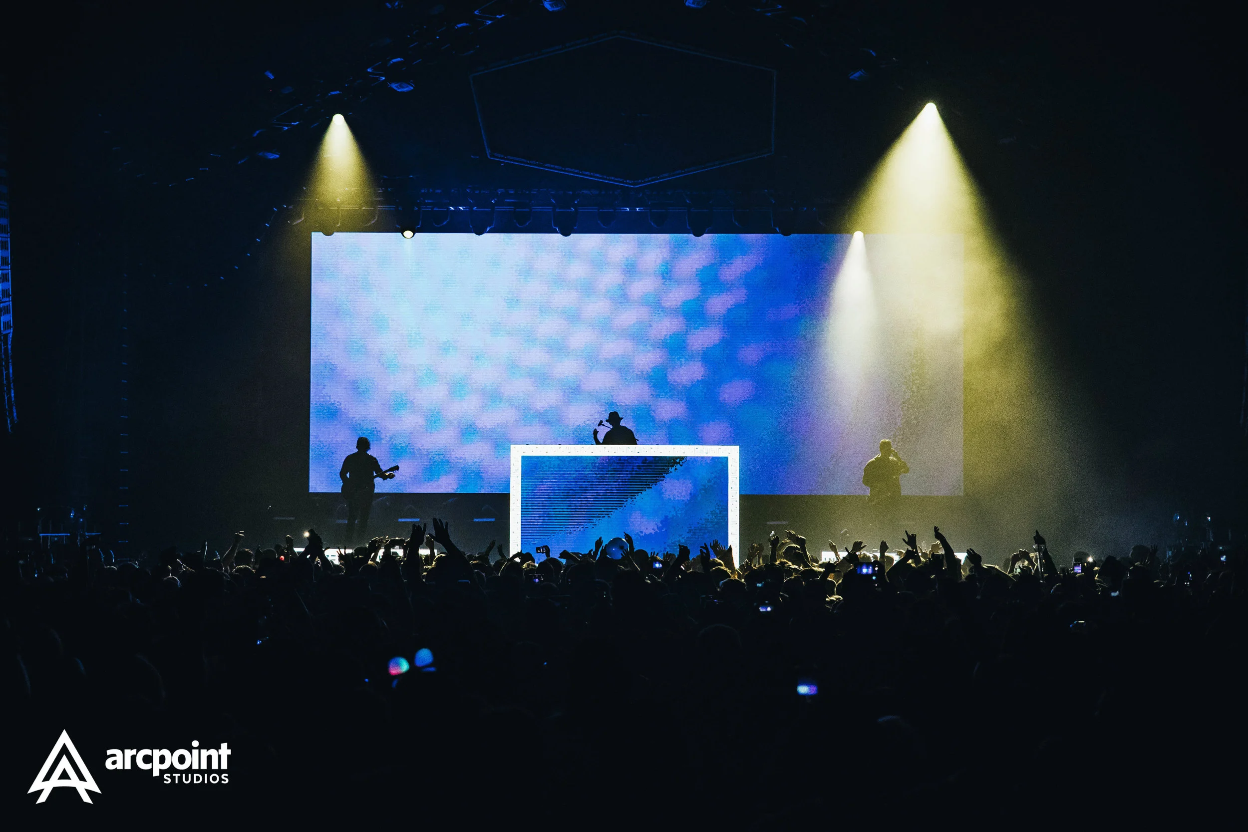 A concert scene with three silhouetted performers on stage, one at a DJ booth with a large digital screen behind them, and the other two holding guitars. The audience is cheering and holding up hands, with some taking photos. Stage lighting creates a