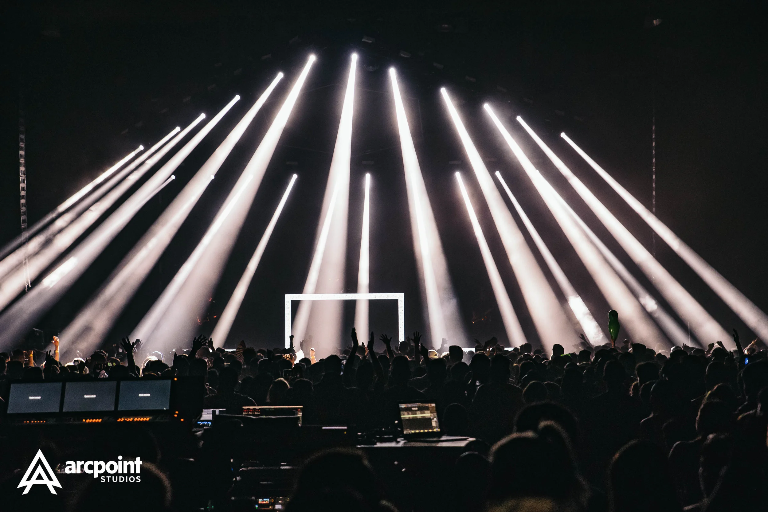 Concert audience with hands raised, stage with bright white spotlights and a rectangular frame.