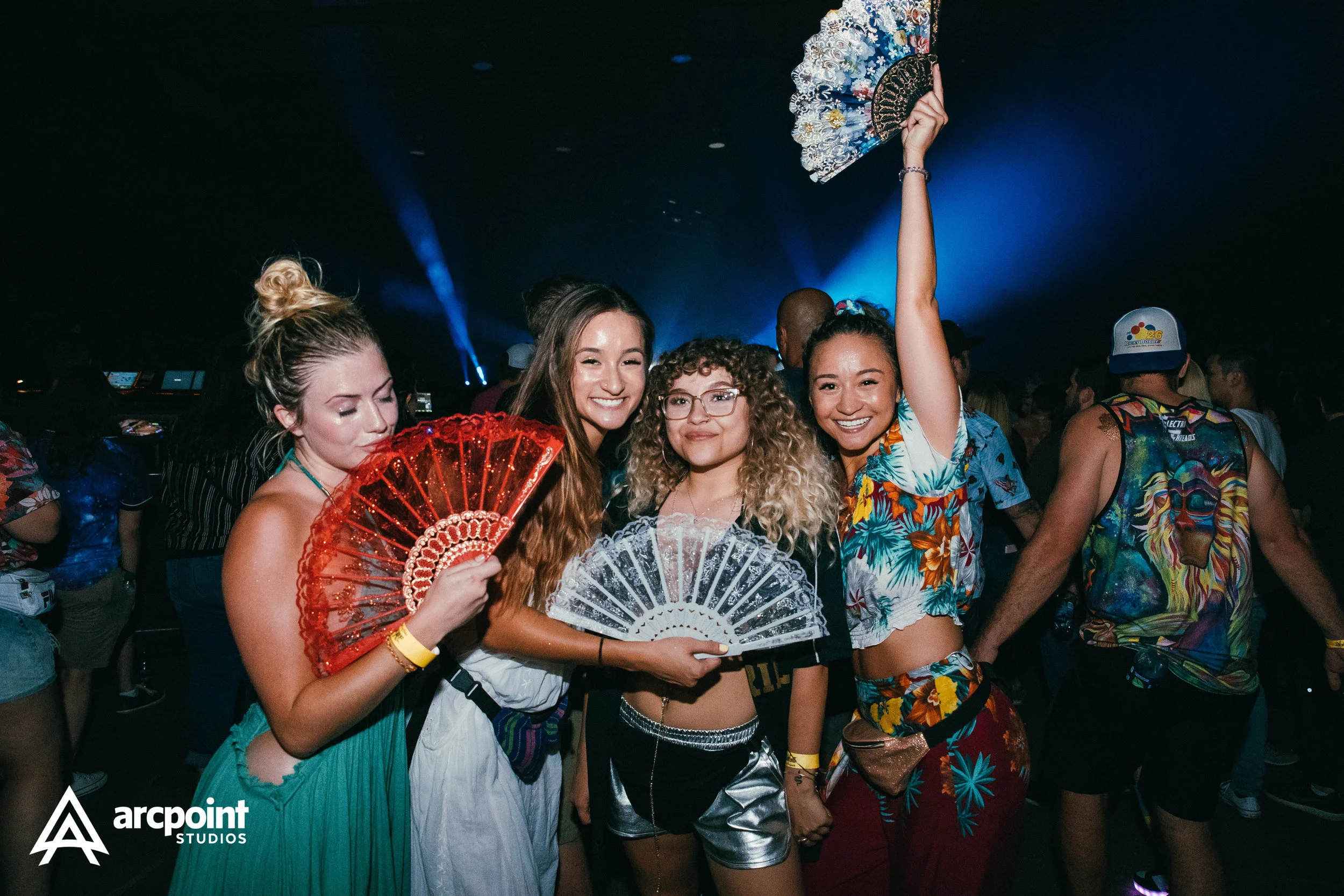 Four young women smiling at a party or concert, three of them holding decorative hand fans, and one woman raising her fan high. The background shows a crowd and stage lighting, indicating a lively event.