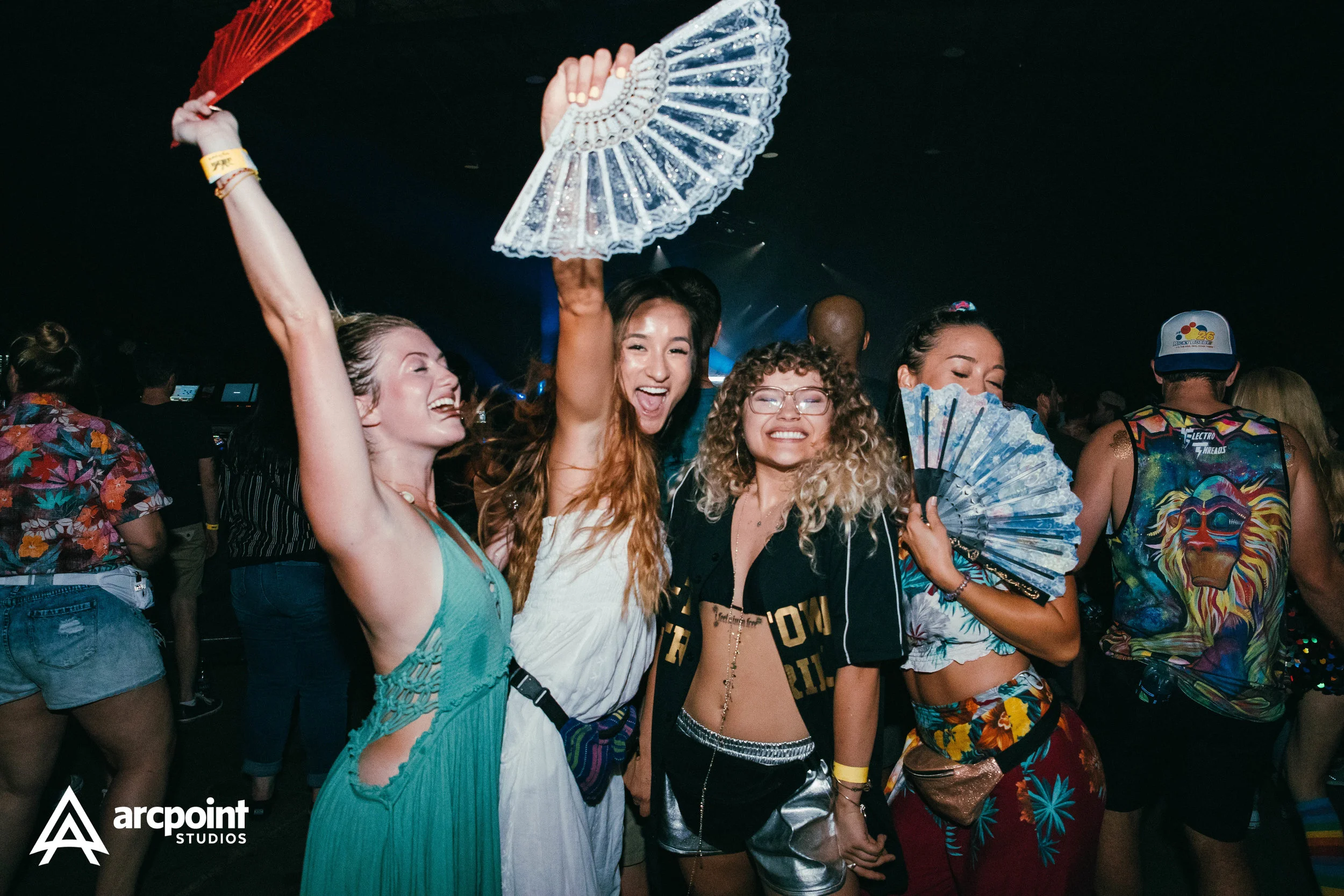 A group of five women enjoying a lively party, holding colorful fans, smiling and having fun in a dark, festive atmosphere.