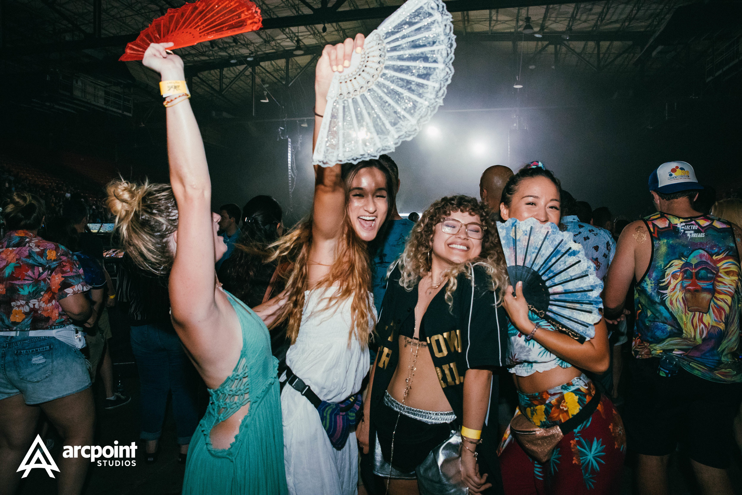 Group of four young women at a lively party, holding decorative fans, smiling, and enjoying themselves in a crowded indoor venue.