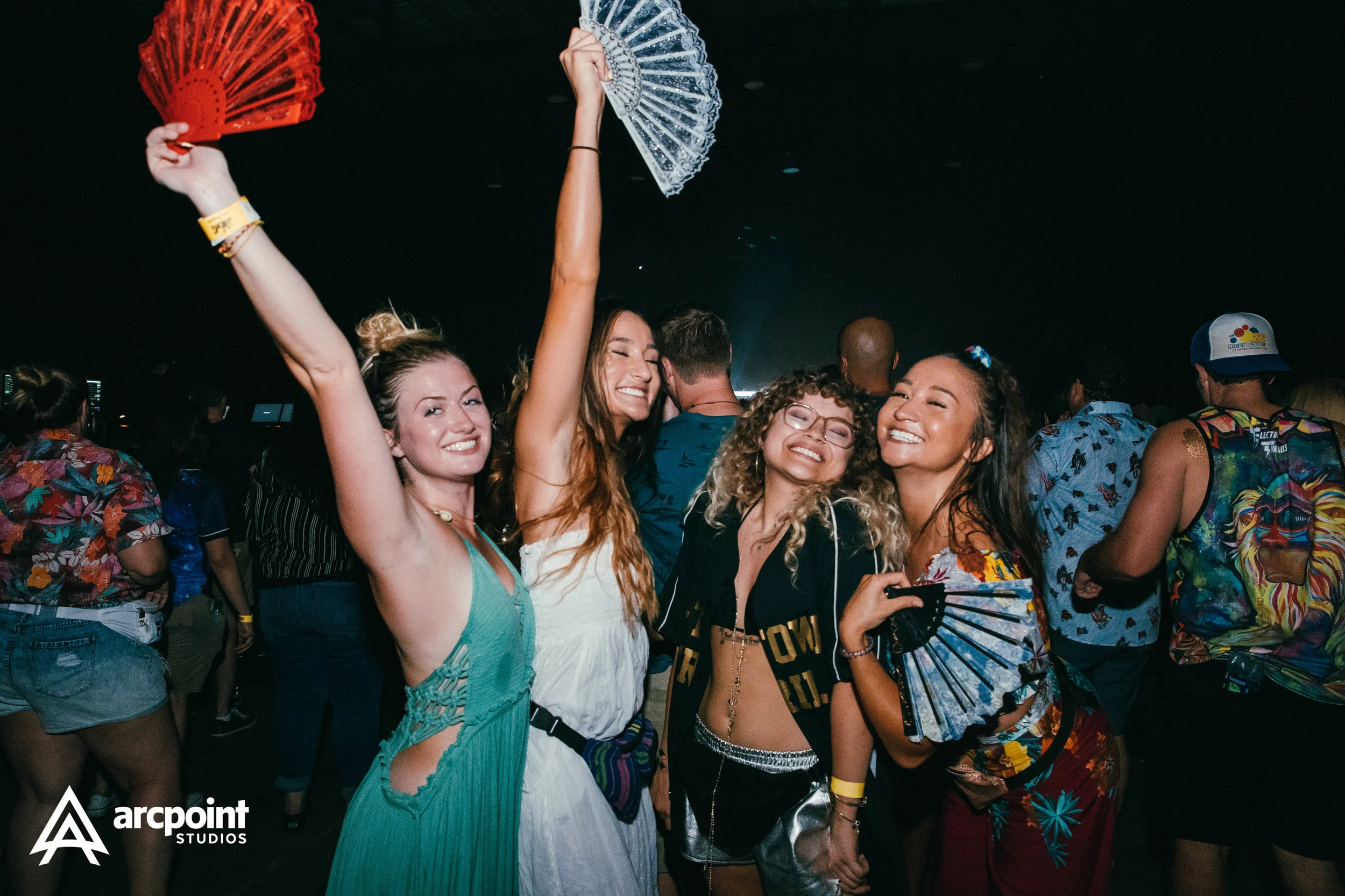 Four young women celebrating at a music festival, smiling and holding hand fans in a dark, crowded environment.