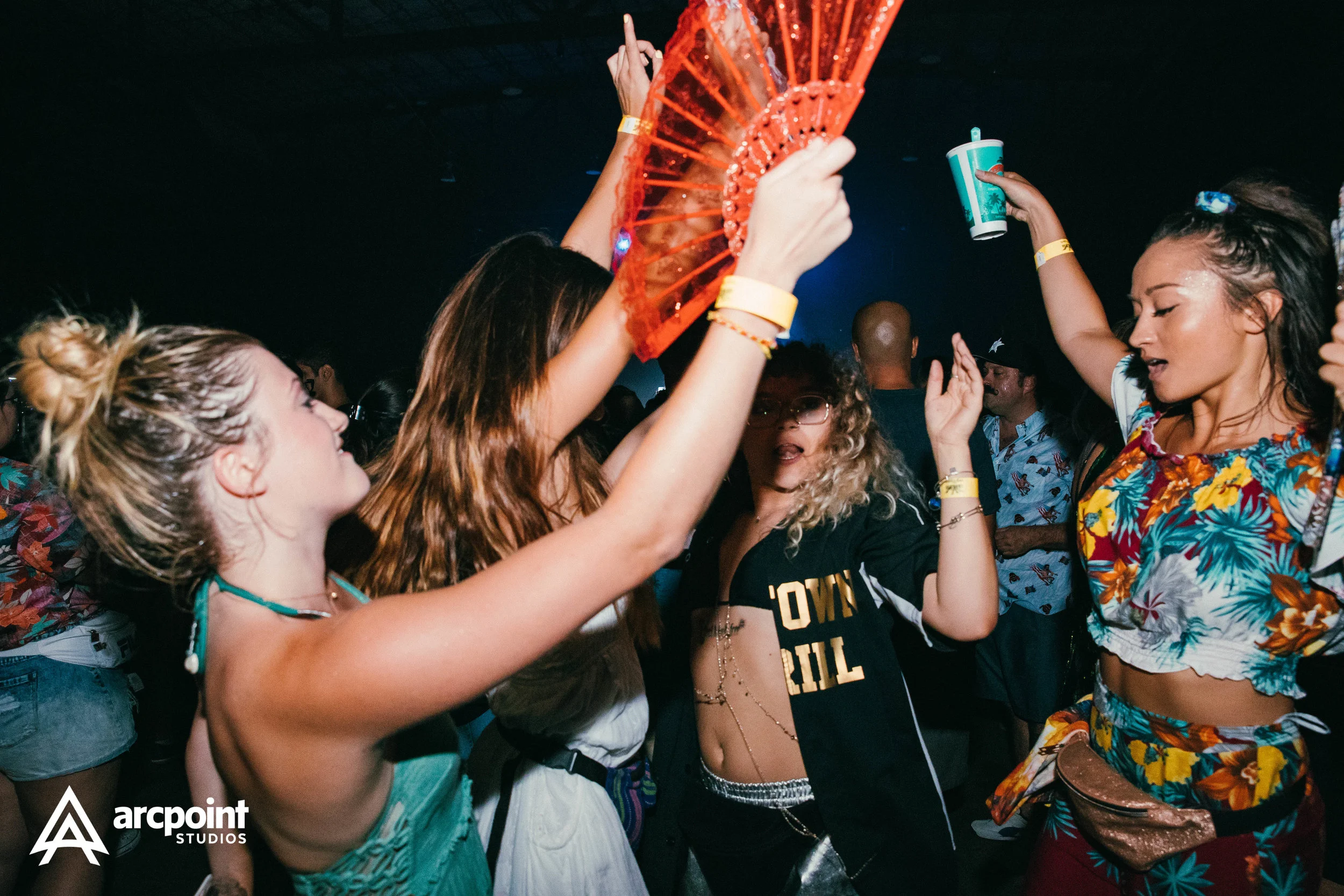 Group of women dancing and enjoying music at a party or club, with some holding drinks and wearing colorful, summer outfits.
