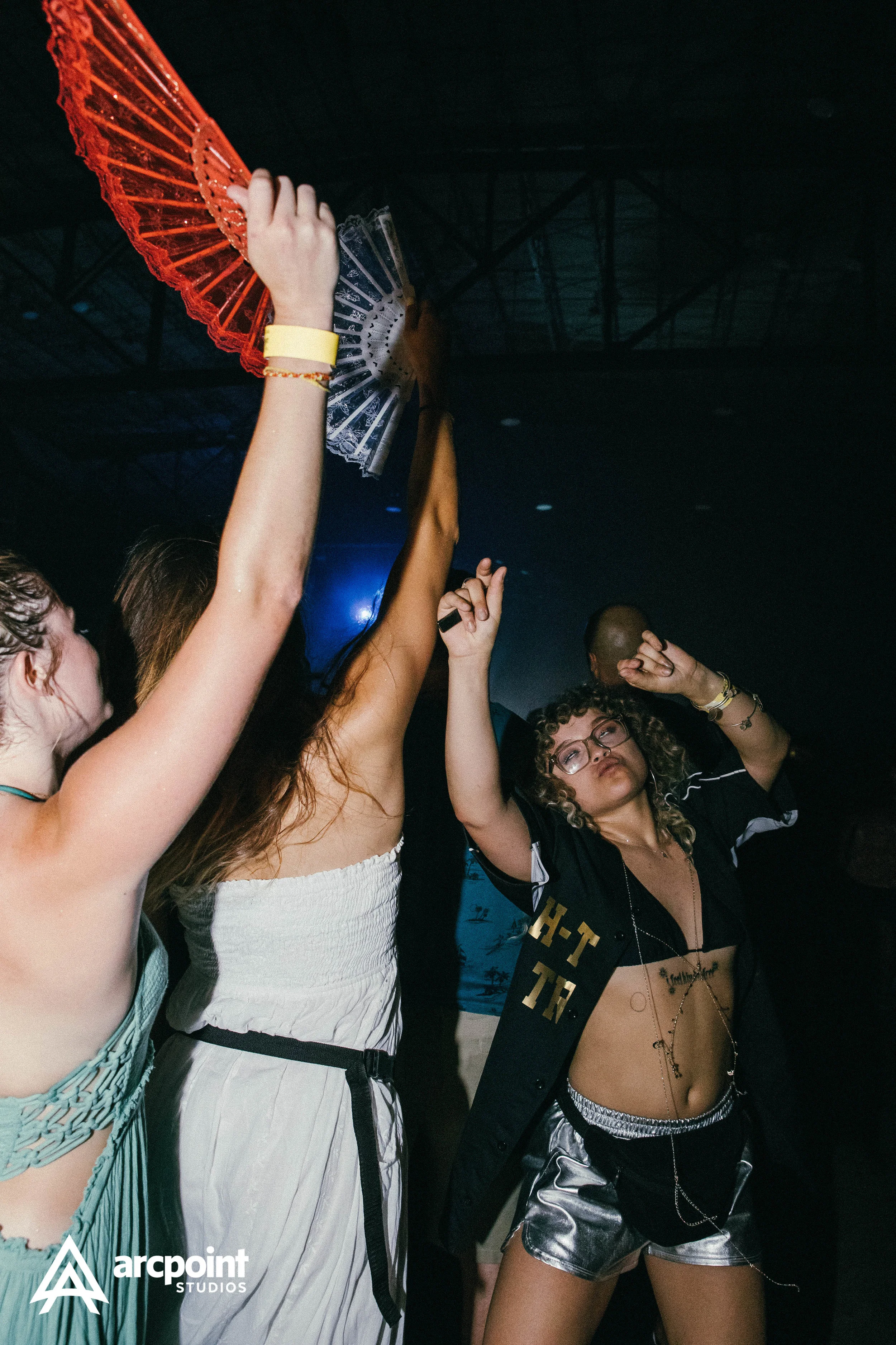 People dancing with raised arms in a dark club setting, colorful fans, and a woman with curly hair, glasses, and jewelry dancing.