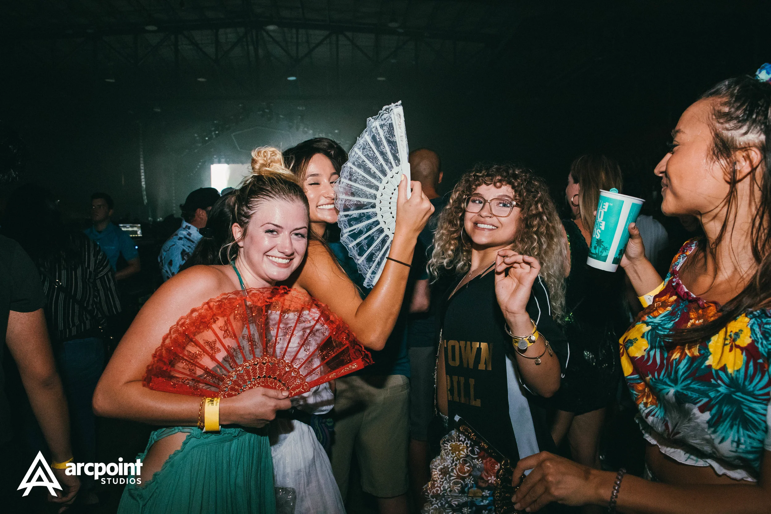Group of women dancing and having fun at an indoor party, holding fans and drinks, with colorful tropical attire and smiling faces.