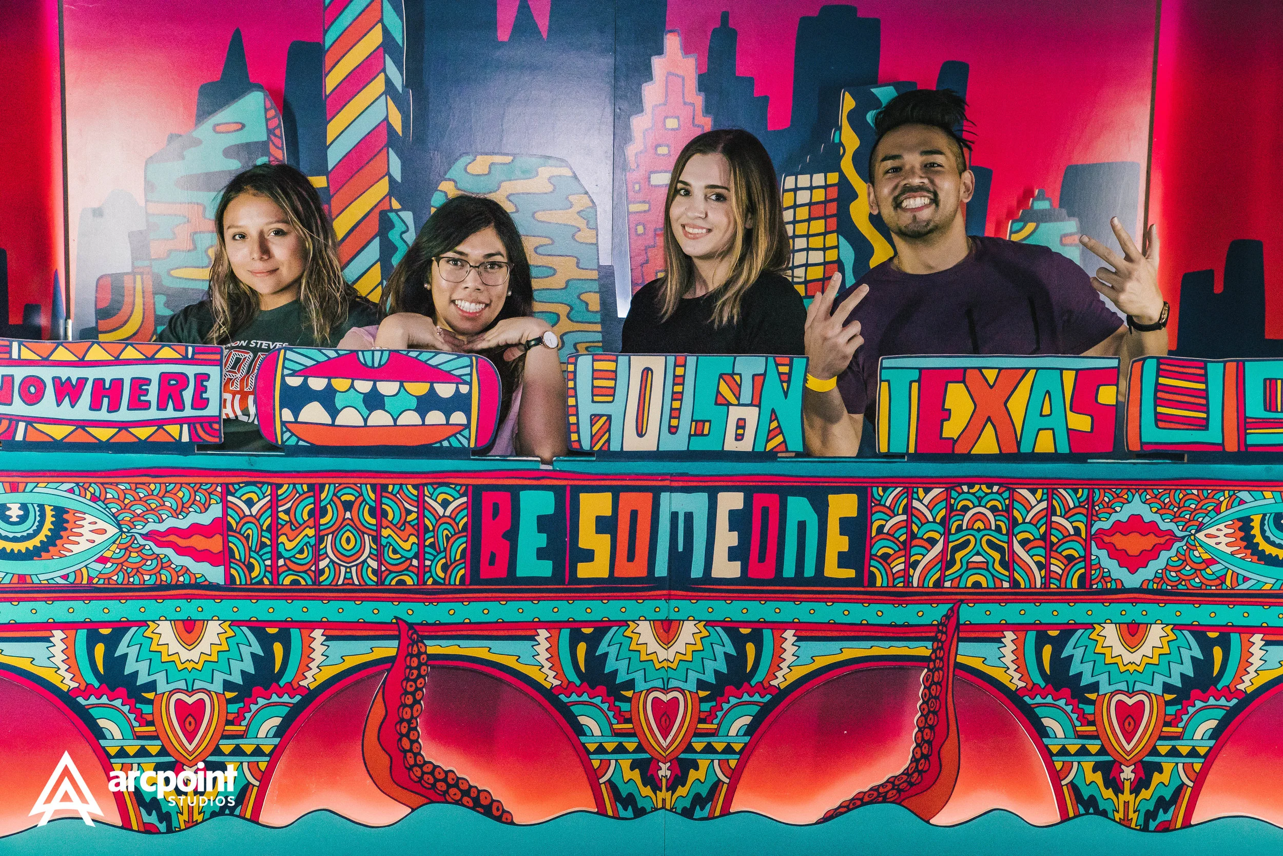 Four young people smiling behind a colorful, graffiti-style bar with signs saying NOWHERE, HOUSTON, TEXAS, and BE SOME ONE, with a cityscape background and decorative patterns, in an indoor arcade or entertainment venue.