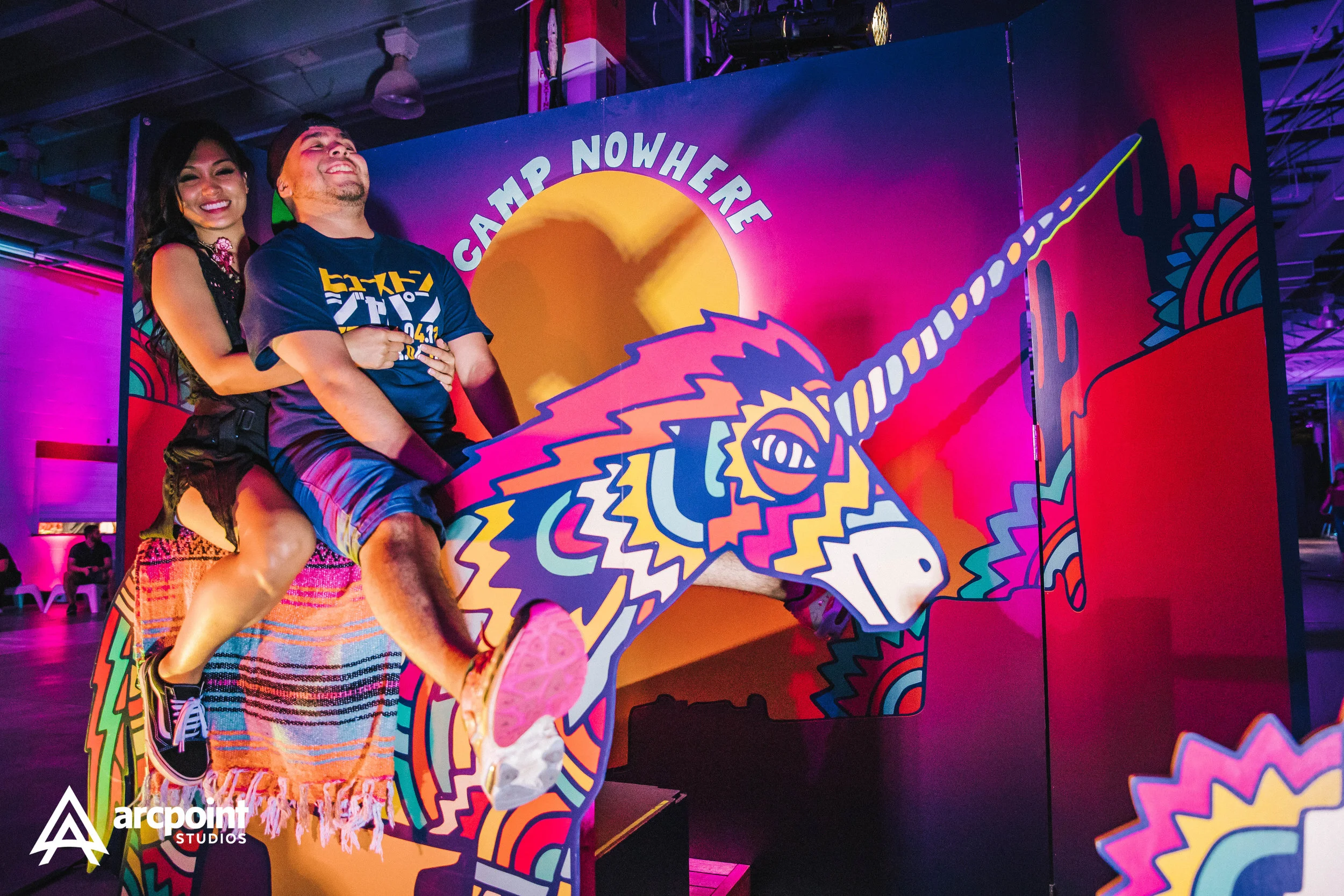 Two people riding a colorful, cartoon-style unicorn on a carnival ride at an indoor event, with a vibrant, illuminated background and a sign that reads "Camp Nowhere."