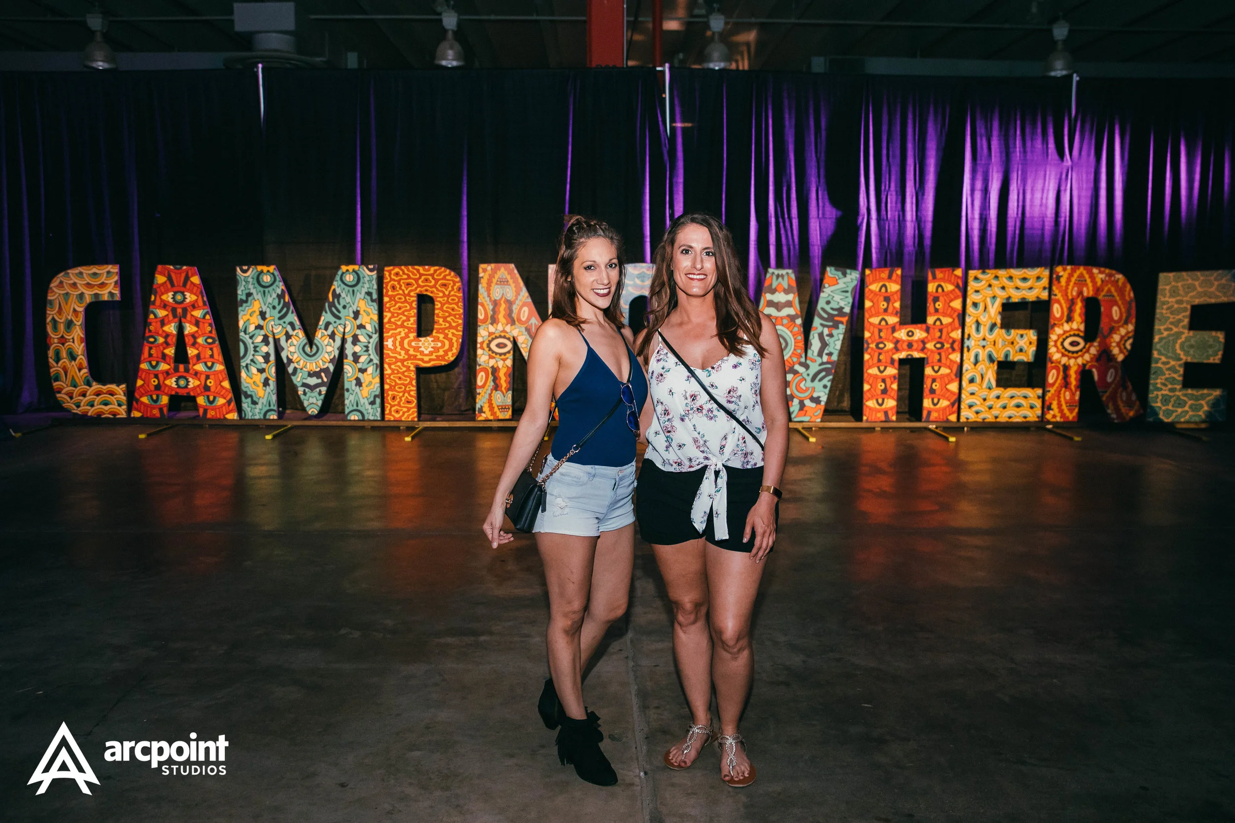 Two women standing side by side in front of large, colorful, patterned letters spelling 'CAMPFIRE' at an indoor event.