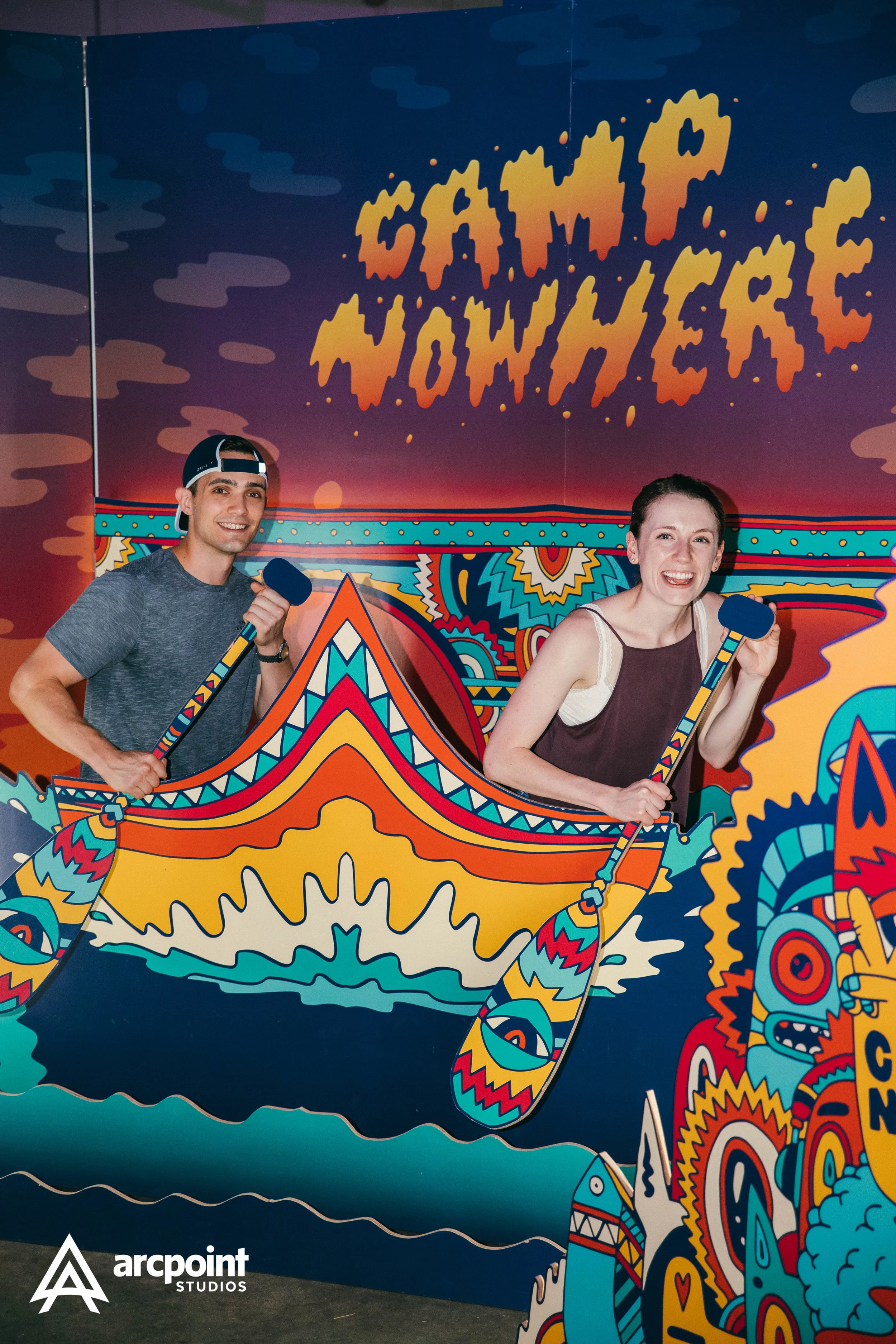 Two people smiling and holding paddles while standing behind a colorful, illustrated boat cutout at a game or amusement attraction, with a vibrant, psychedelic background and the words 'Game On' in bold orange and yellow letters.