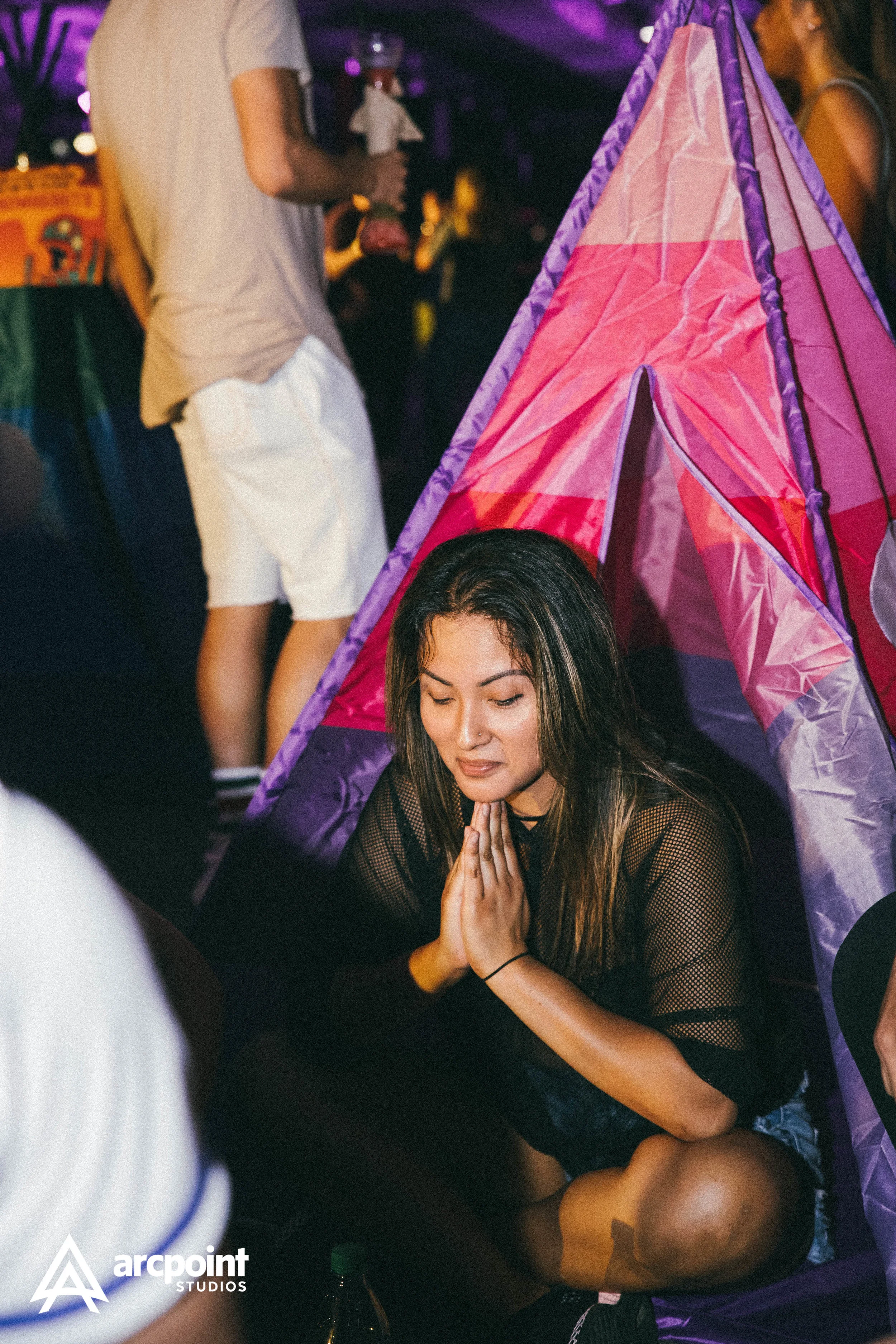 A woman sitting cross-legged and praying inside a pink and purple pop-up tent at a party. People are visible in the background.