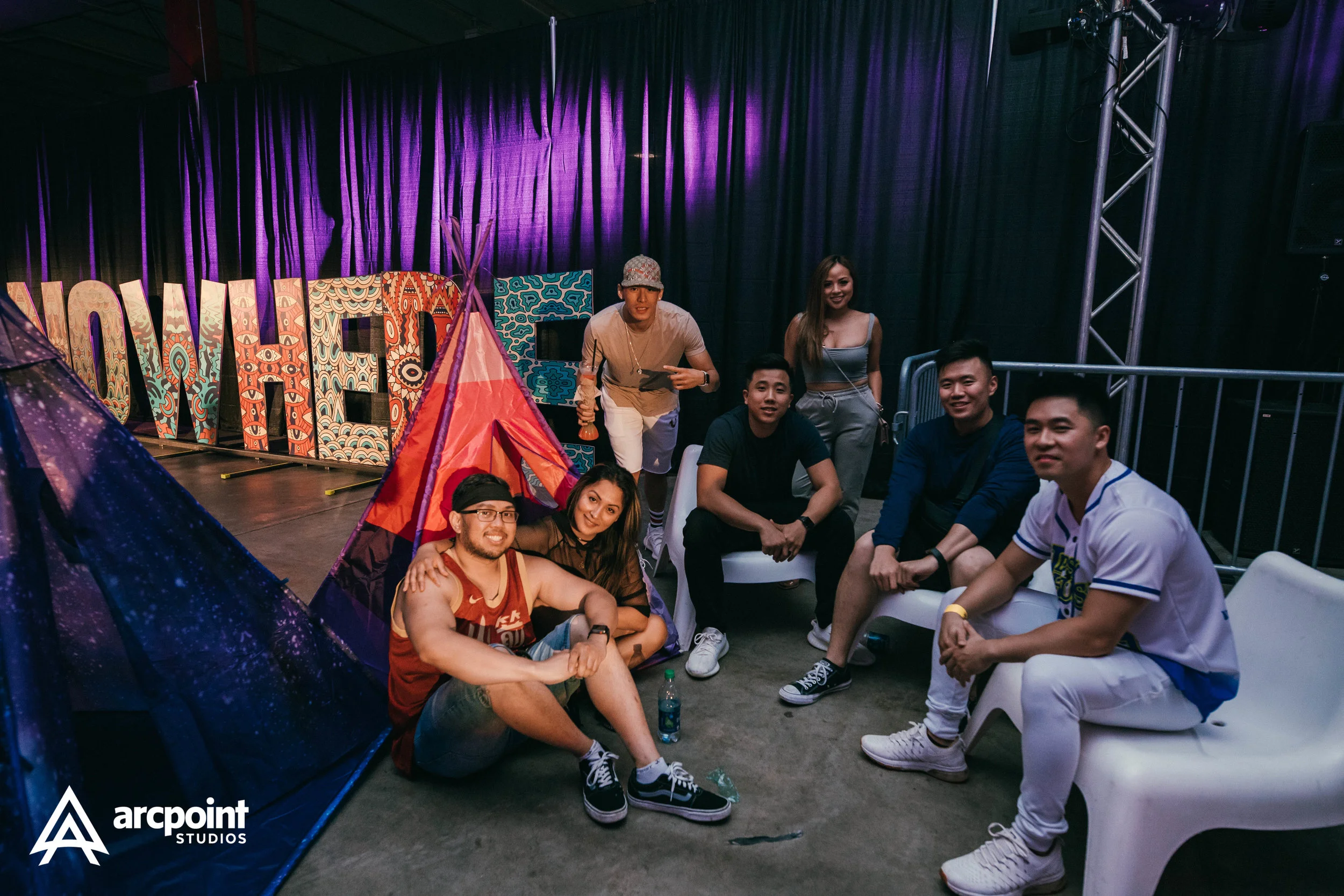 Group of friends posing in a room with colorful tent and creative lettering background, with a purple curtain and stage lighting.