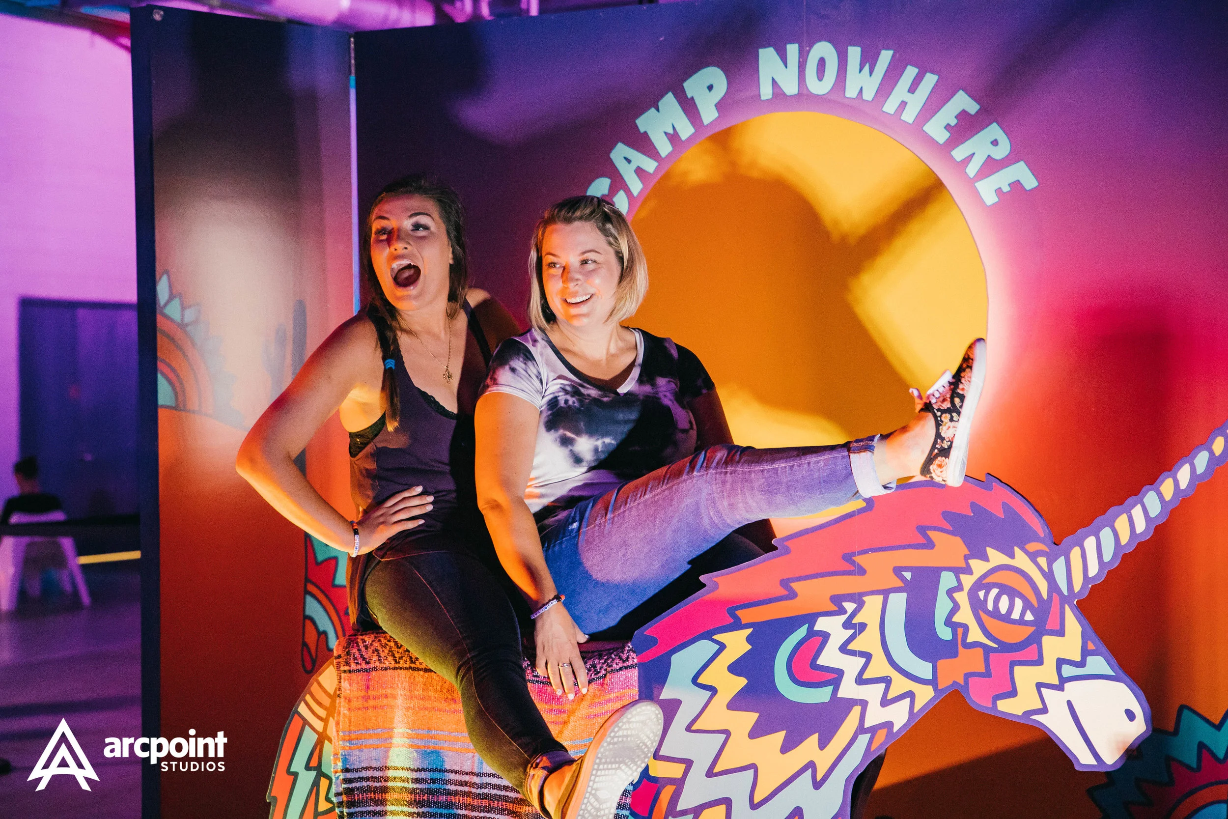 Two women riding a colorful, stylized unicorn at a themed attraction with a sign reading "Camp Nowhere" in the background.