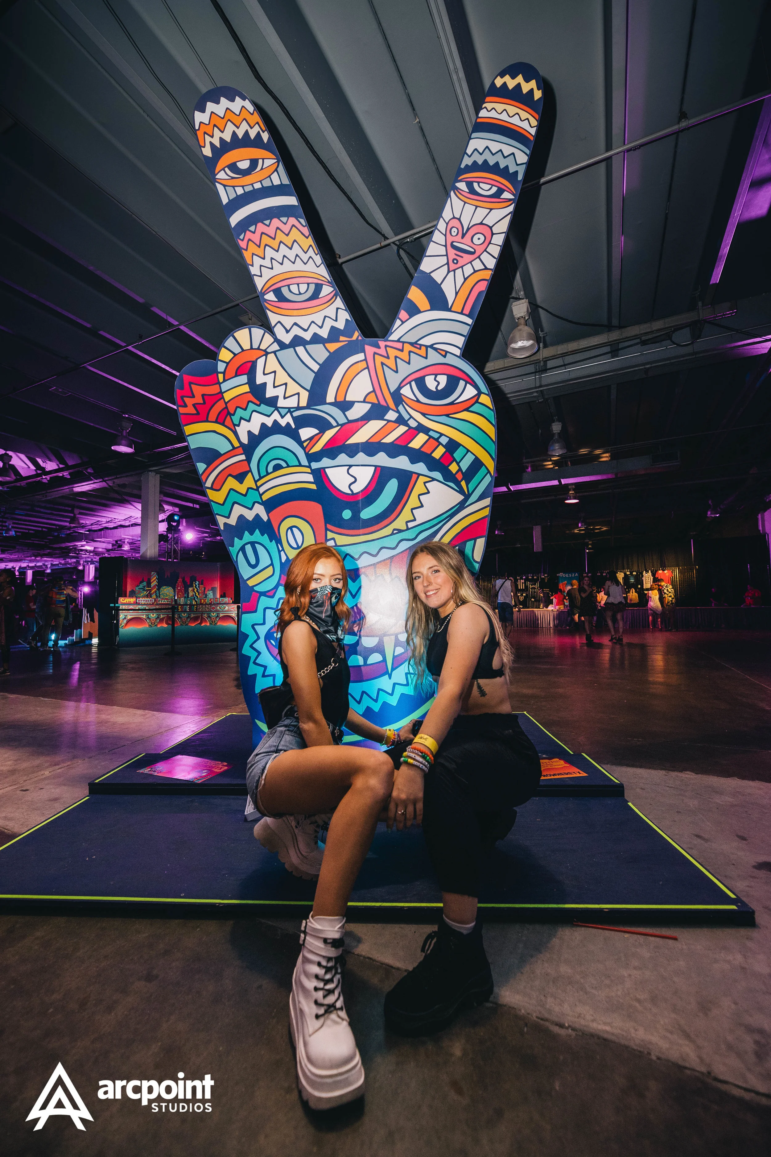 Two young women squatting in front of a large, colorful artwork of a peace sign with a face, eyes, and a tongue, displayed indoors at an event venue with purple lighting.