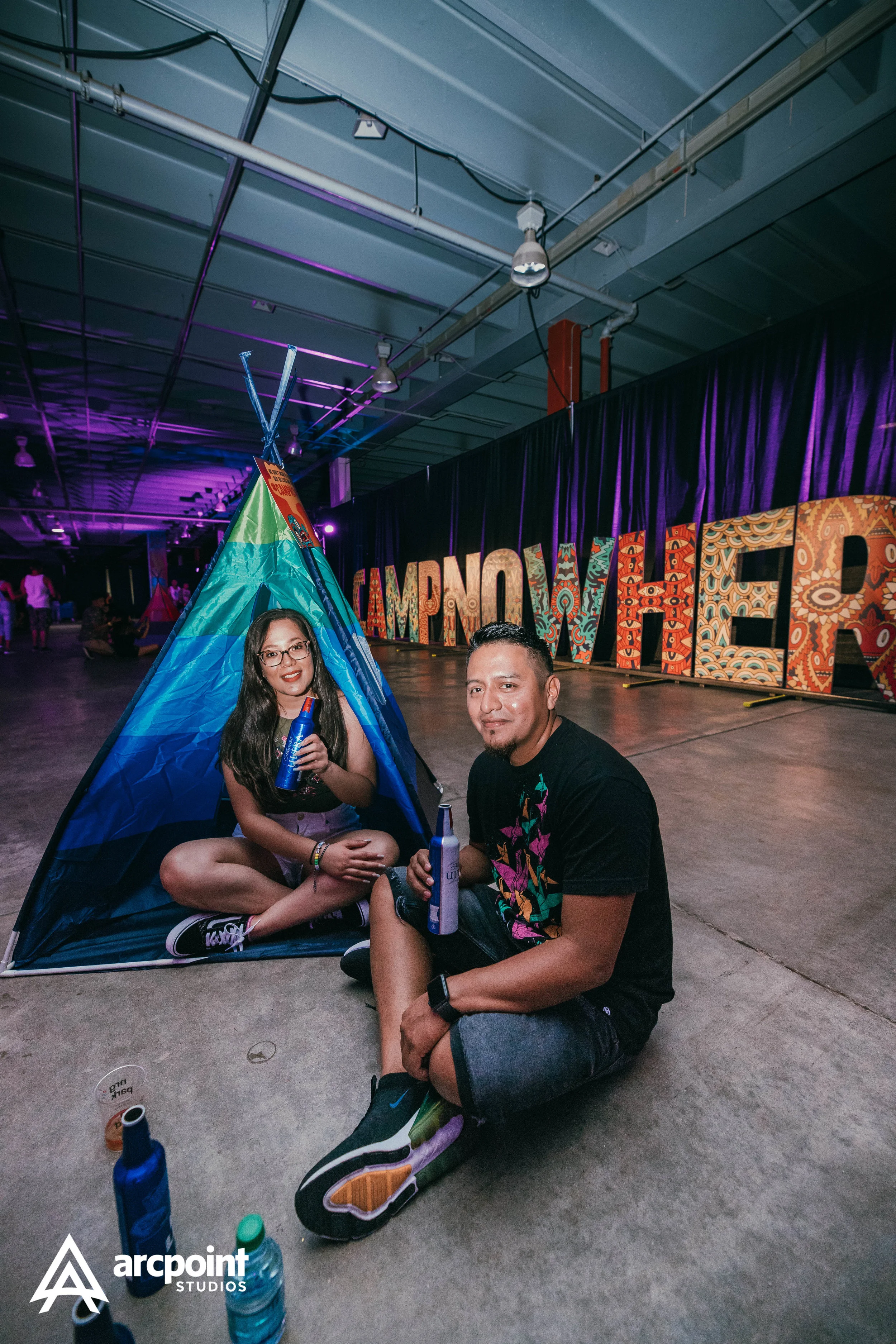 Two people sitting on the floor in front of a camping tent, holding blue bottles at an indoor event with colorful sign letters spelling 'IMPONT' in the background.