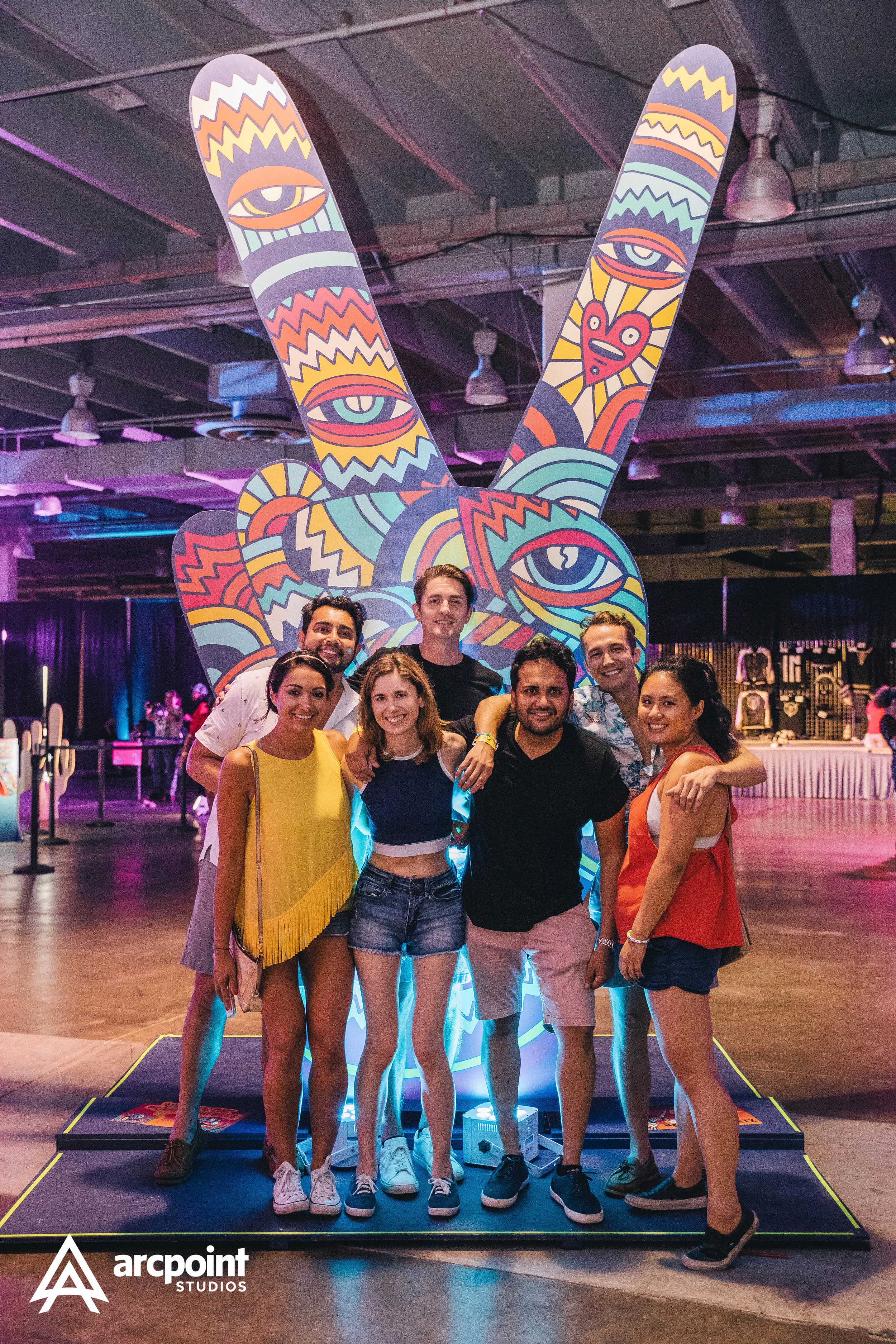 Group of six friends posing together at an art exhibit featuring a large colorful sculpture of a peace hand sign with eye and face designs, in an indoor gallery space with dim lighting and vibrant purple and pink accents.