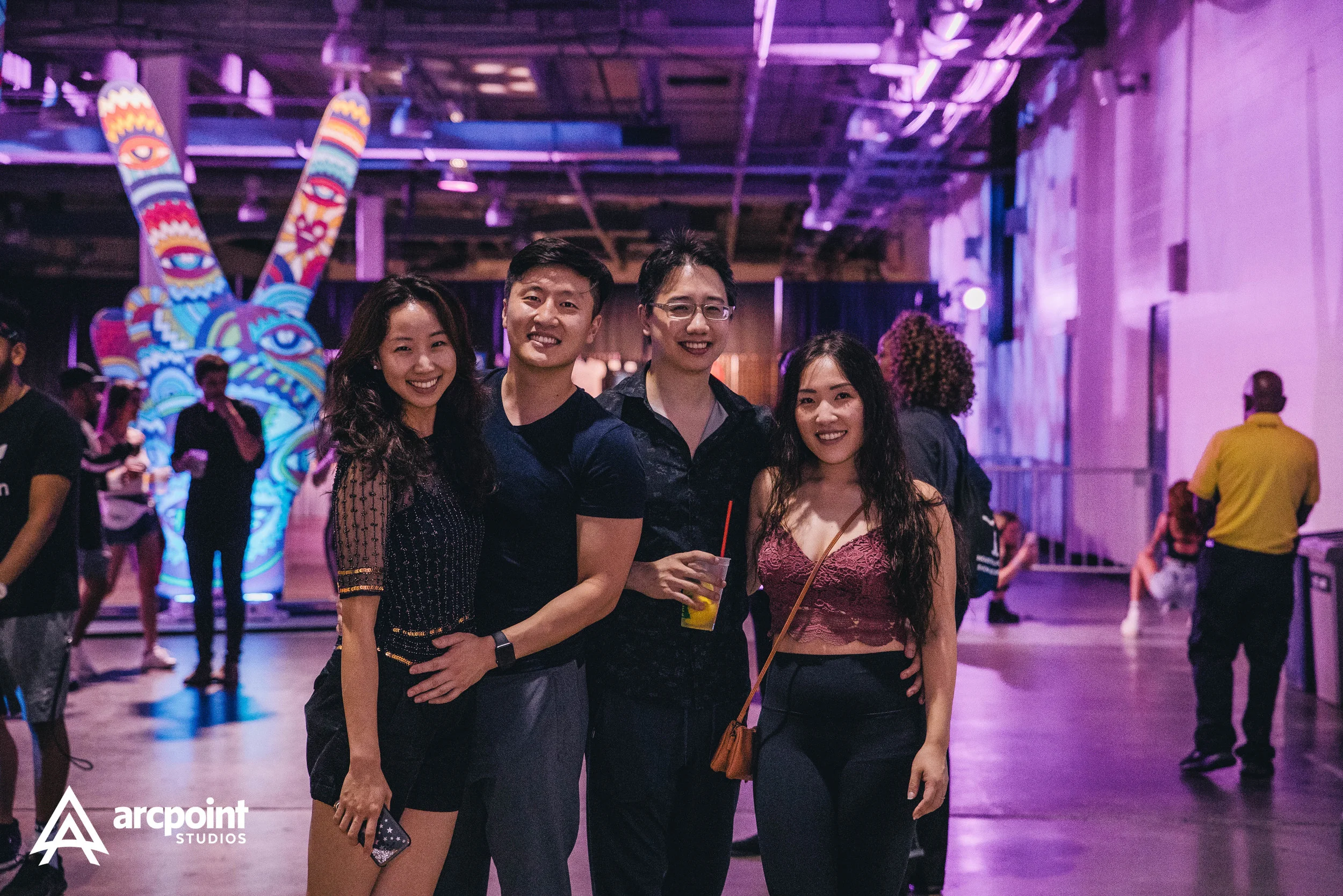 Four young adults, two women and two men, smiling and posing together at an indoor event with colorful lighting and art installation in the background.