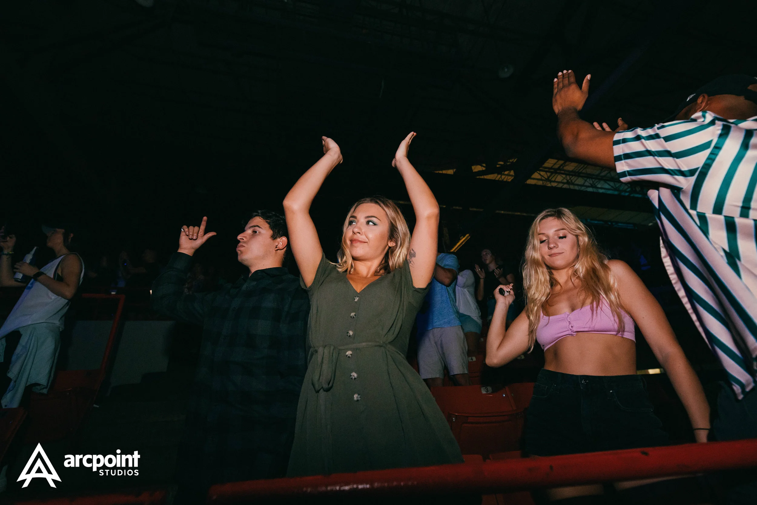 Group of young people dancing and enjoying music at a concert or nightclub, with some raising their hands and eyes closed.