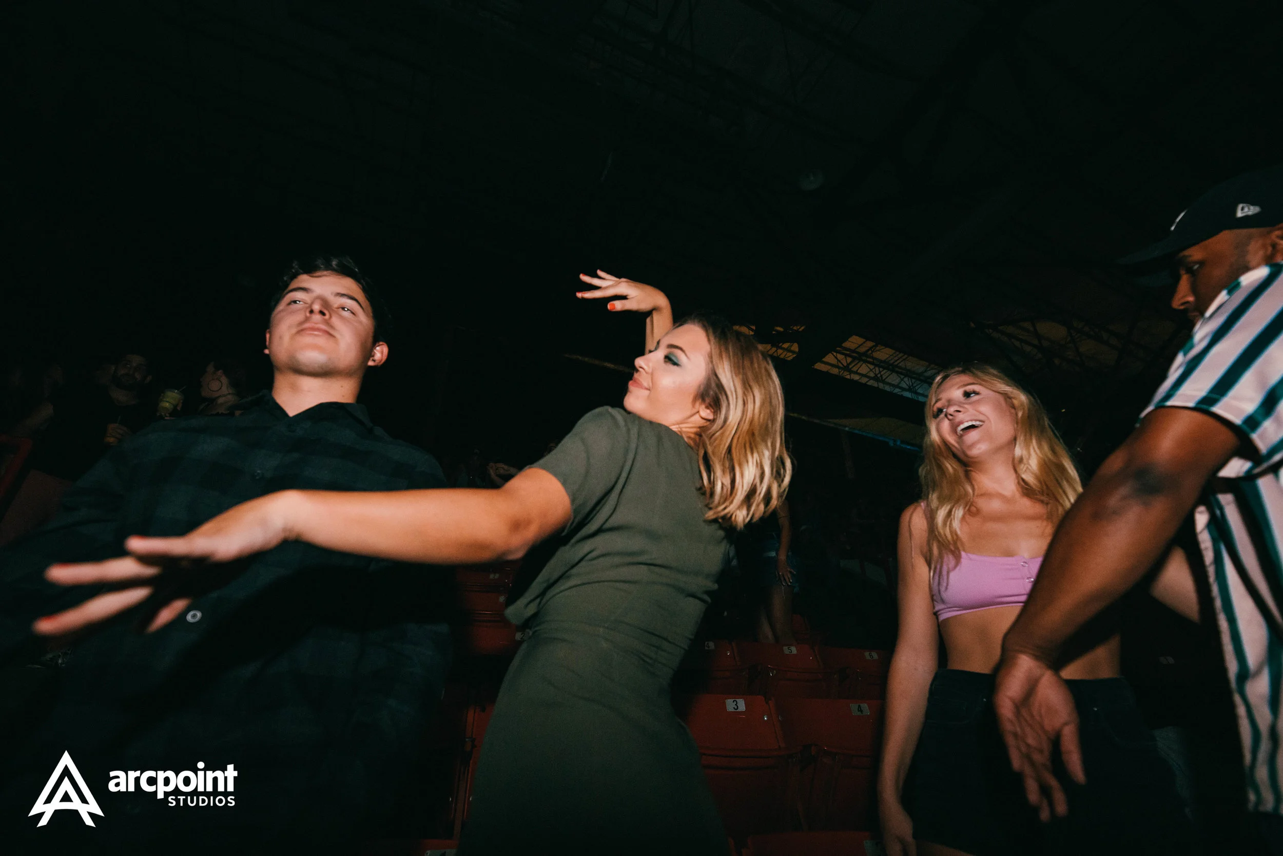 People dancing and enjoying themselves at an indoor event. The scene appears lively and fun, with one woman in a green dress striking a dance pose, a woman in a pink top smiling, and a man in a striped shirt engaging with them.